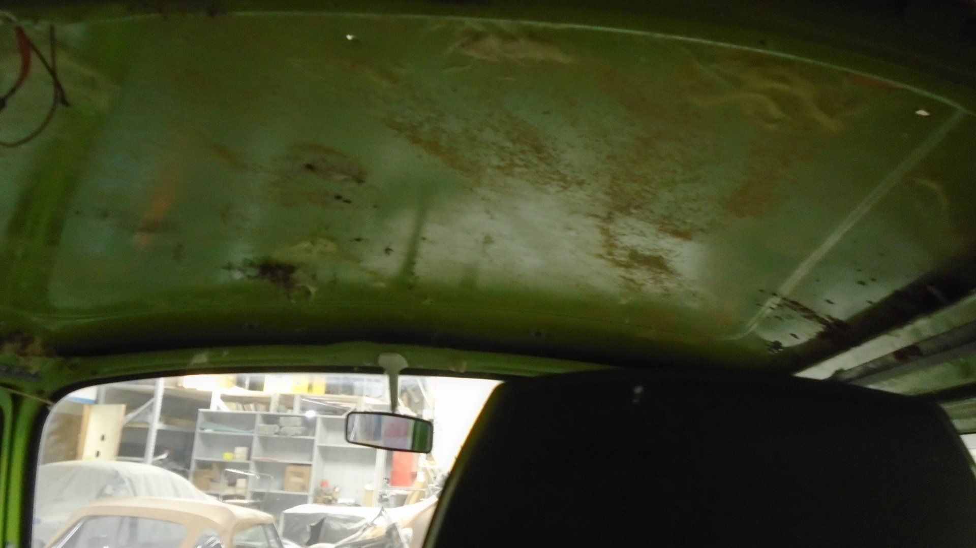 Green vehicle interior ceiling with rust, a rearview mirror, and an obscured black hood.