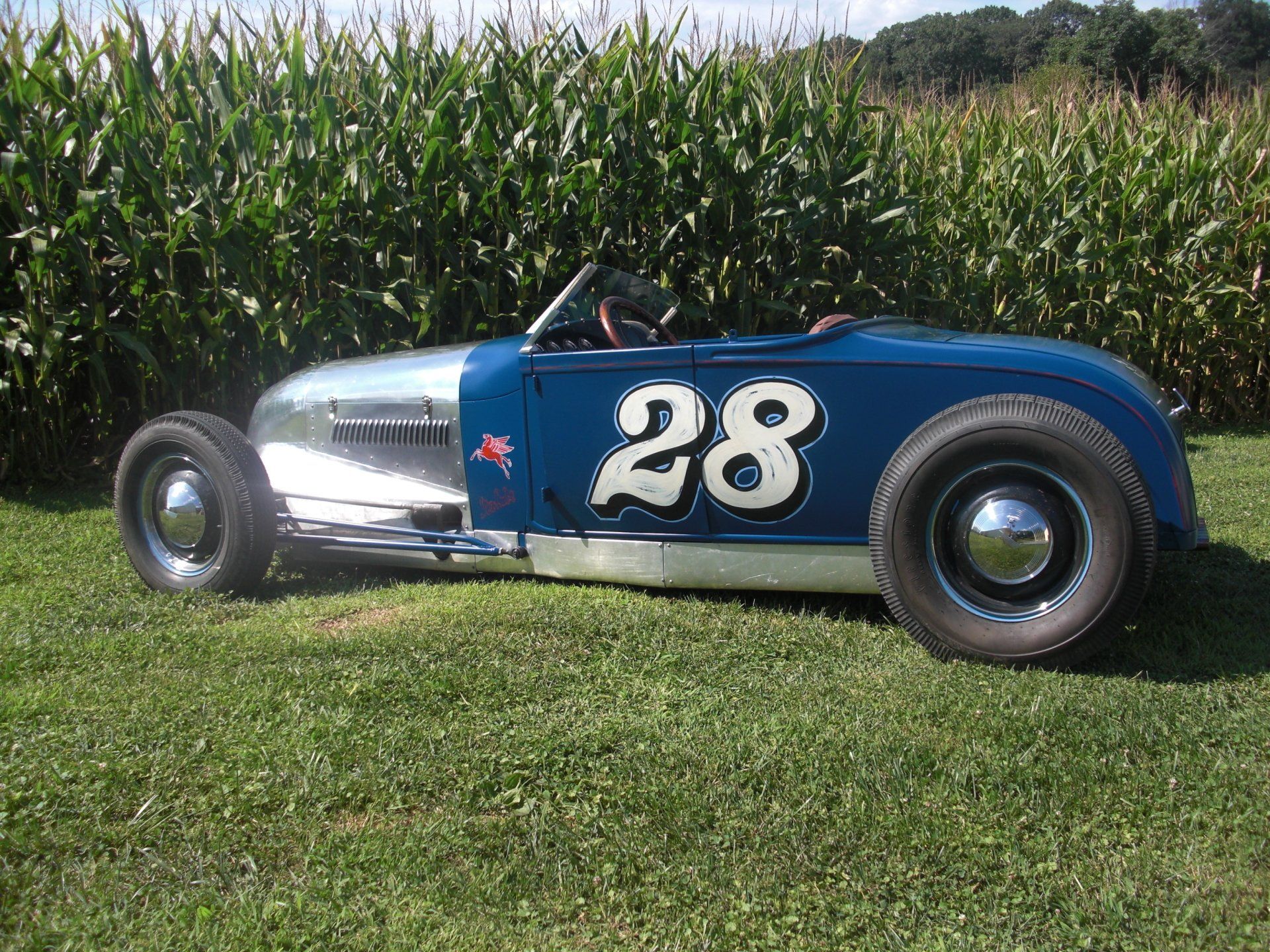 Blue and silver vintage race car with number 28 parked on grass, cornfield in the background.