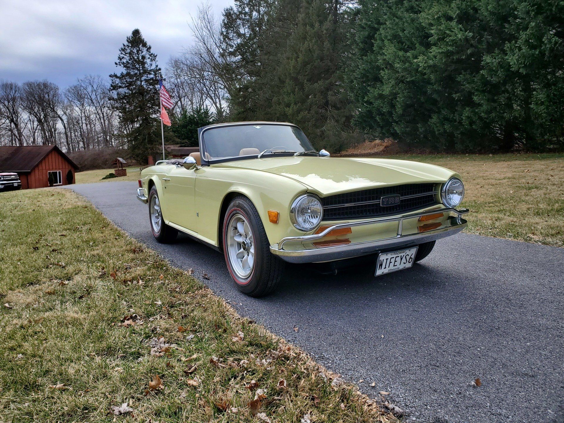 Yellow Triumph TR6 convertible parked on a driveway with a small red building and trees in the background.