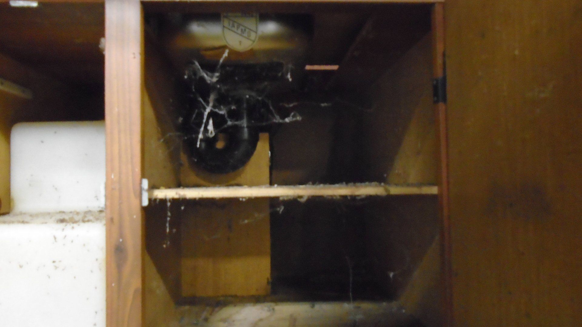 Inside of a wooden cabinet with a dark object behind a wood block, covered in cobwebs.