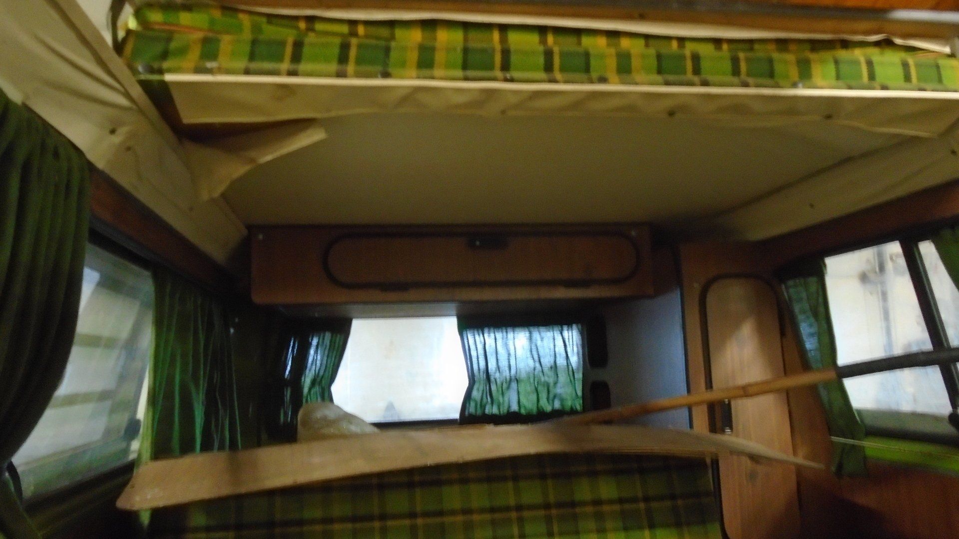 Interior of a vintage camper van with green plaid bedding and curtains, overhead storage.
