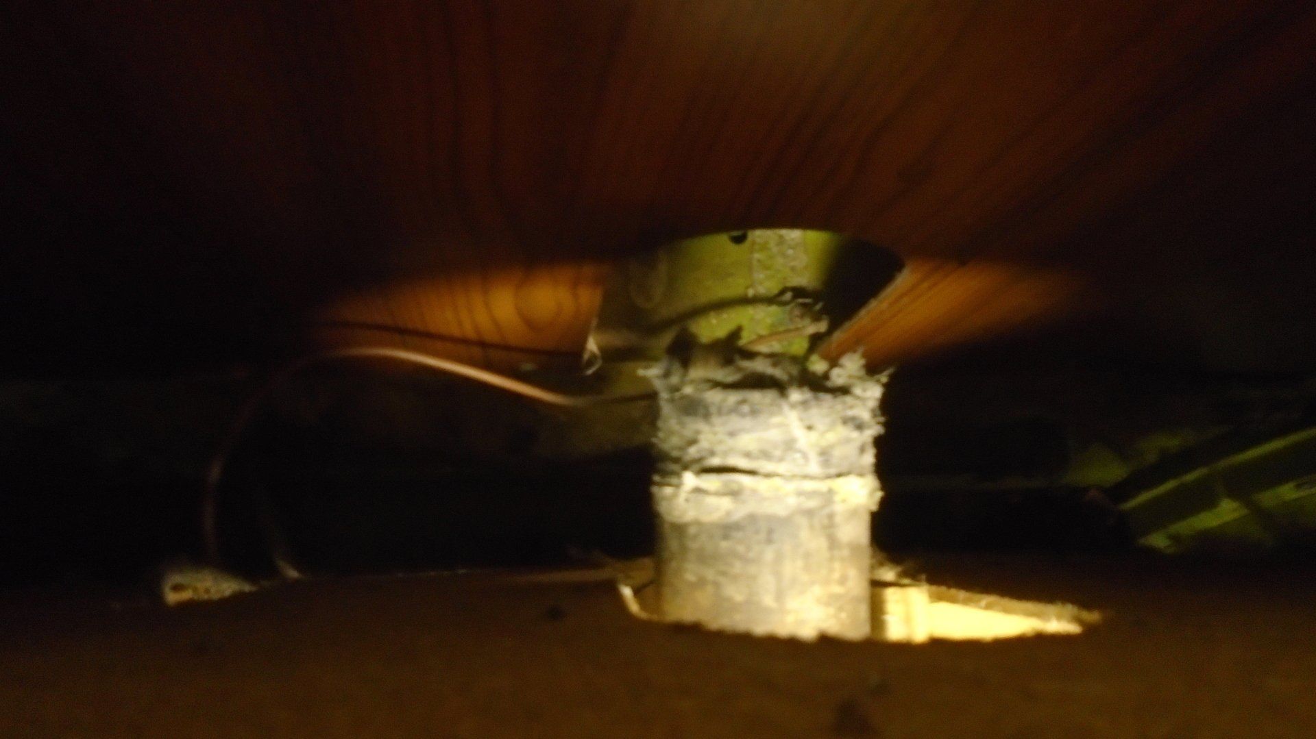 A vertical metal pipe passes through a wooden floor. The pipe has insulation and is dark.