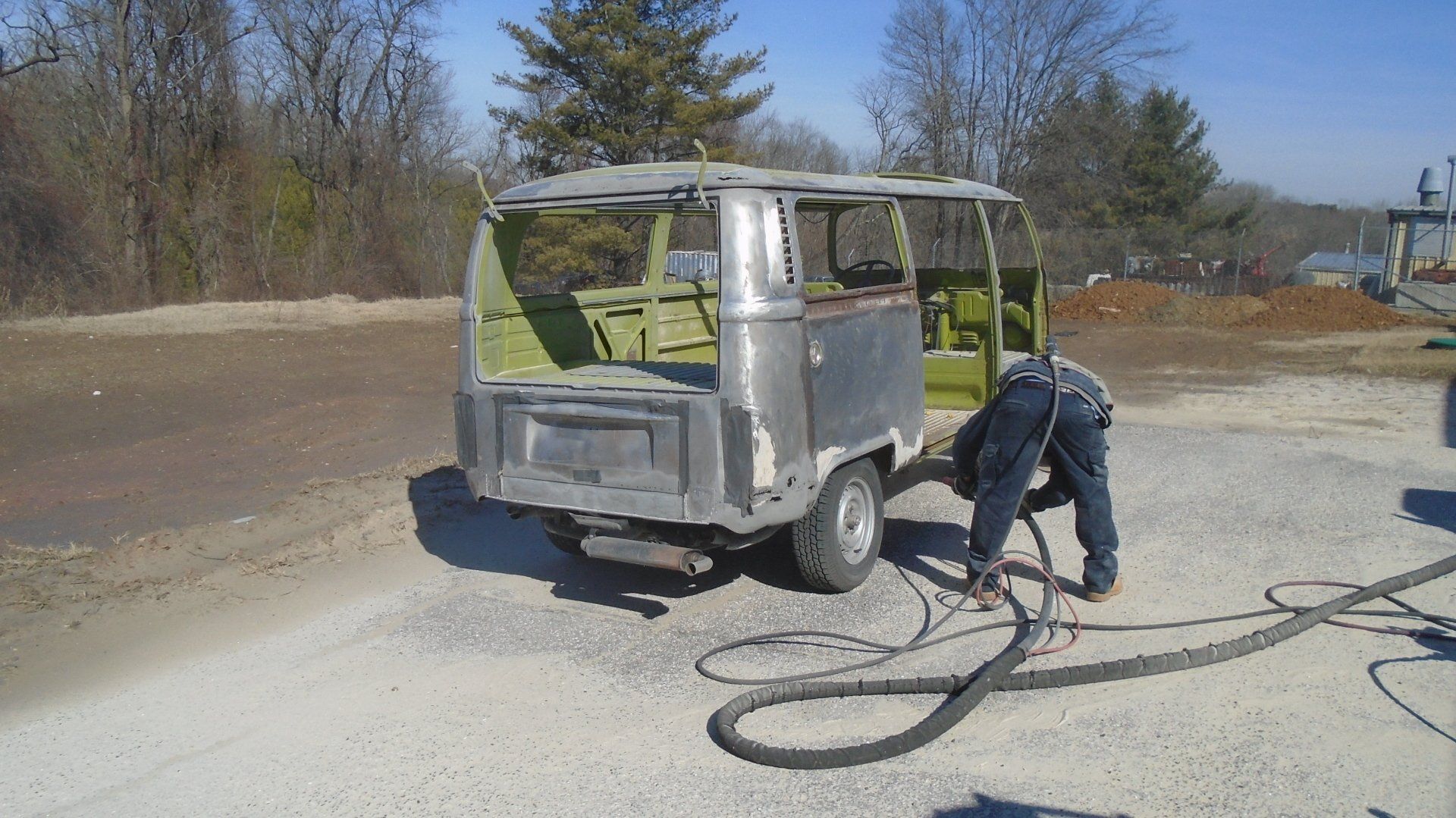 A person sandblasting a vintage van, stripped of its paint. Outdoor setting, sunny day.