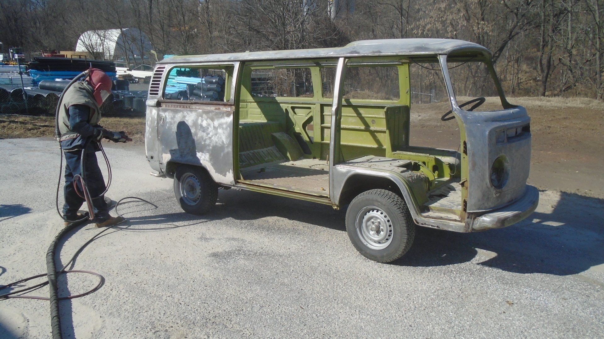 A person sandblasting a vintage Volkswagen bus outdoors. The bus is stripped to metal, with some green paint visible.