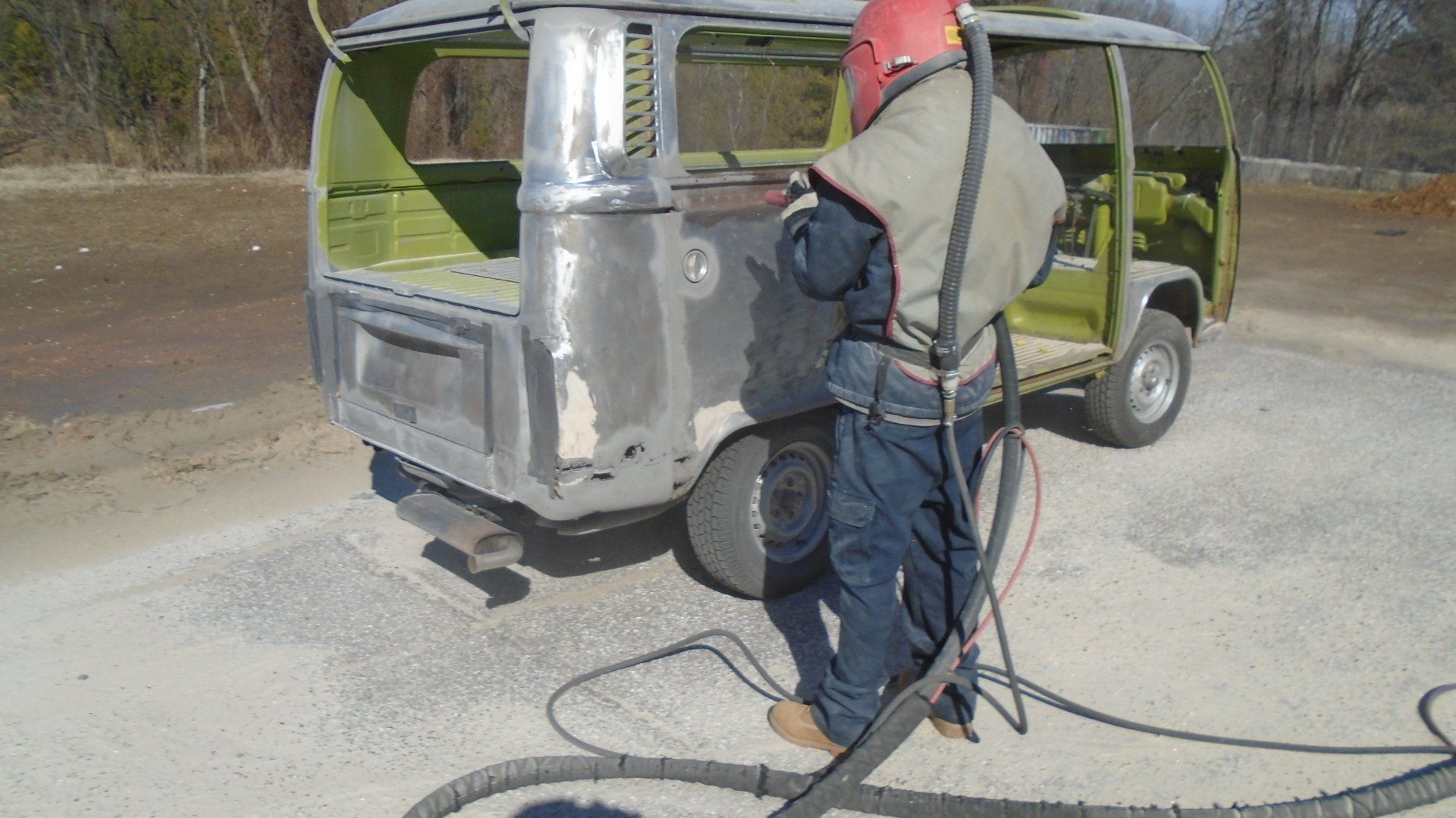 Person sandblasting a vintage van outdoors, wearing protective gear. Gray and green colors are prominent.