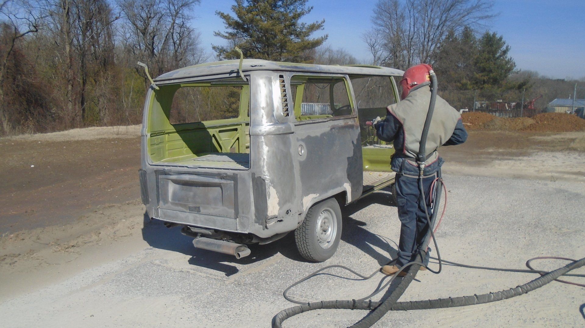 Man sandblasting a vintage Volkswagen van, outdoors. The van is partially disassembled and painted.