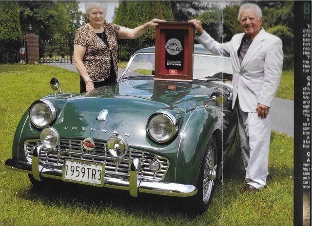 Couple with a classic green Triumph TR3, holding an award.  They stand in front of their car on grass.