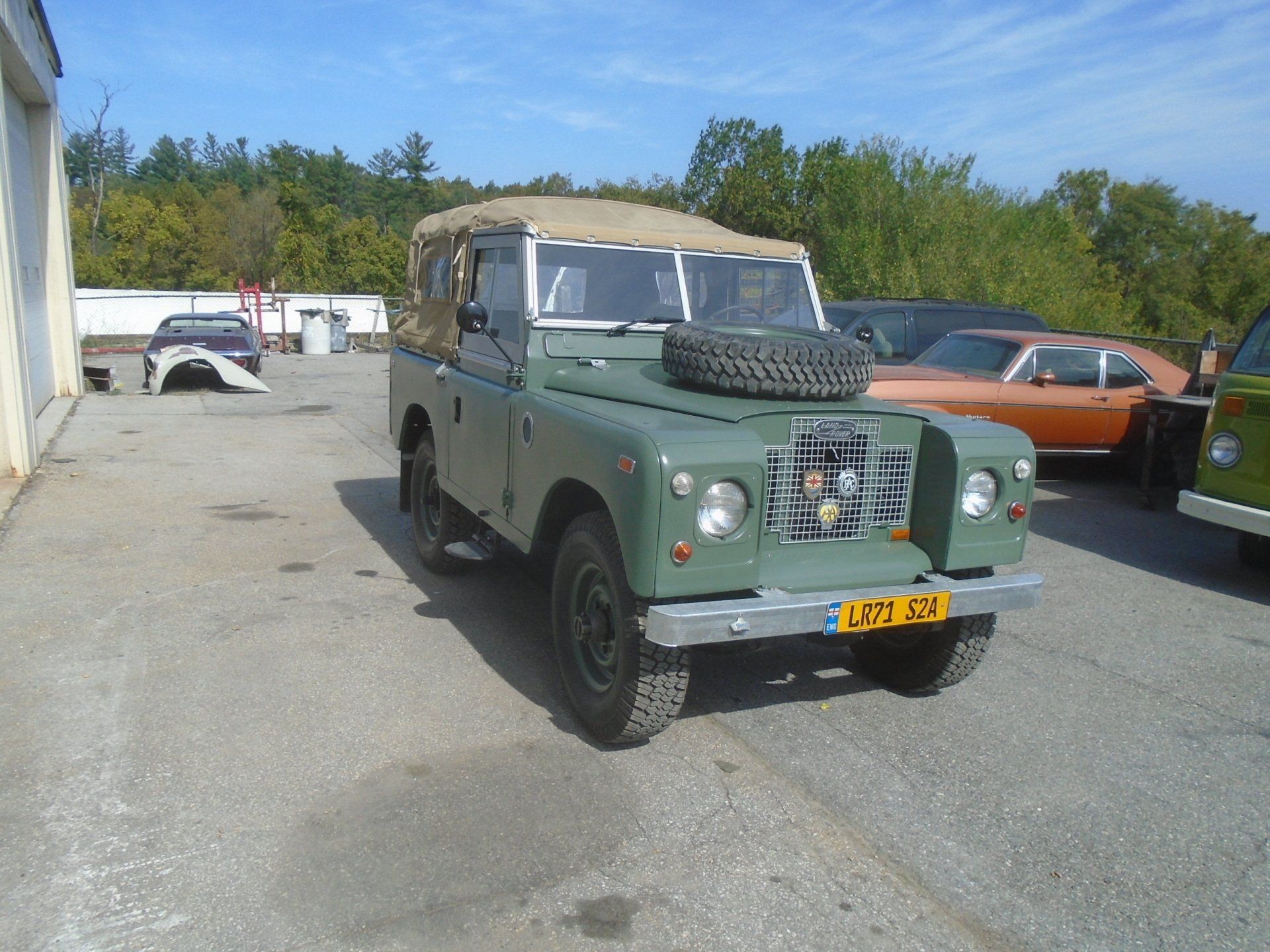 Green vintage Land Rover with a tan roof, parked outside near other vehicles.
