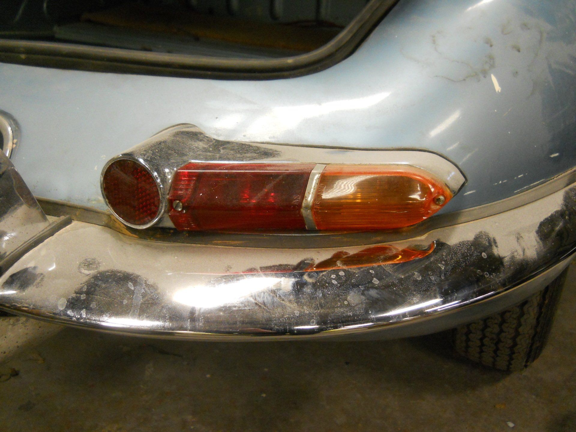 Close-up of a vintage car's rear, showing taillight and chrome bumper; light blue body, red and amber lens.