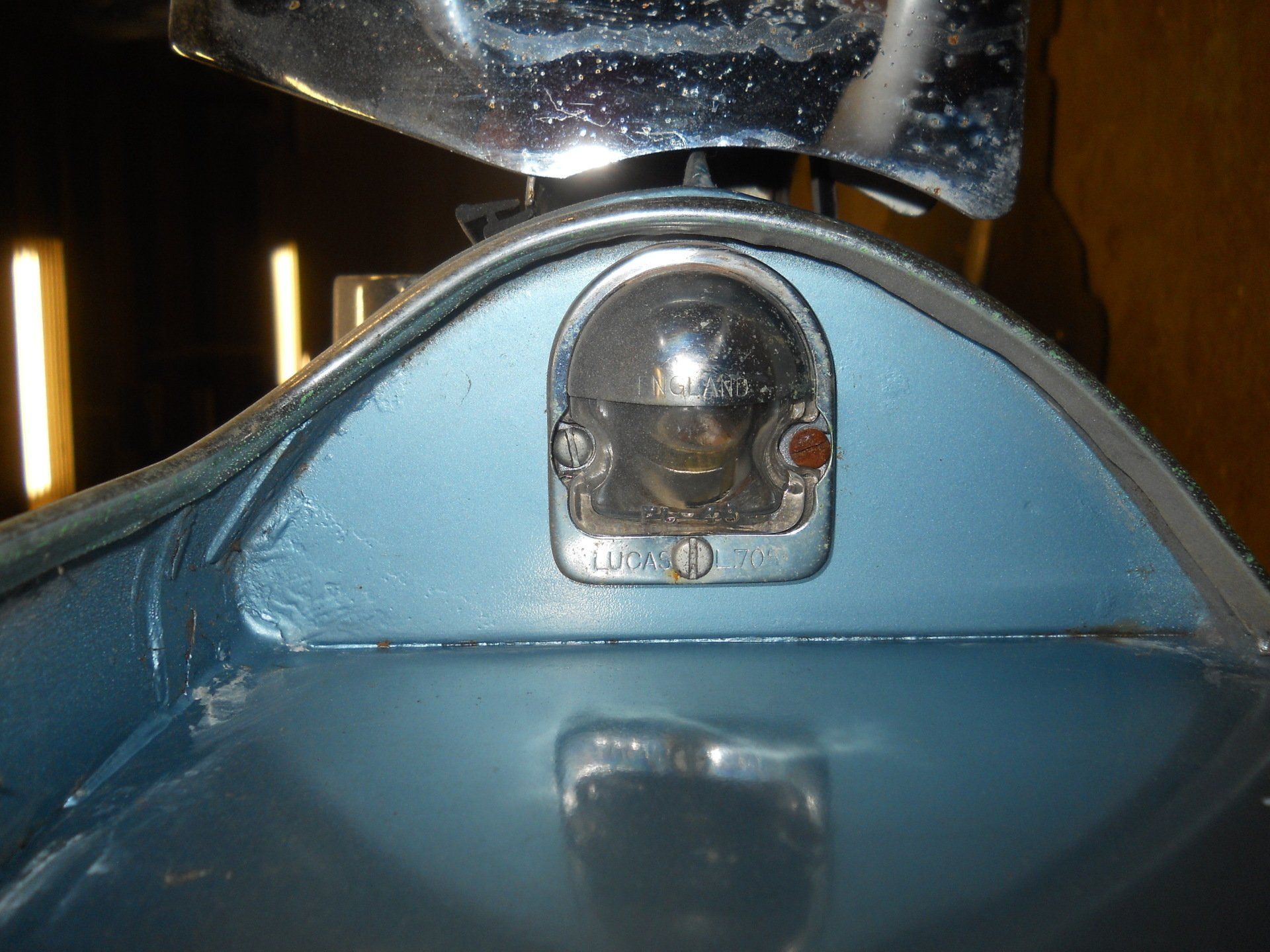 Blue car trunk detail with a small, rounded taillight. Chrome bumper above.