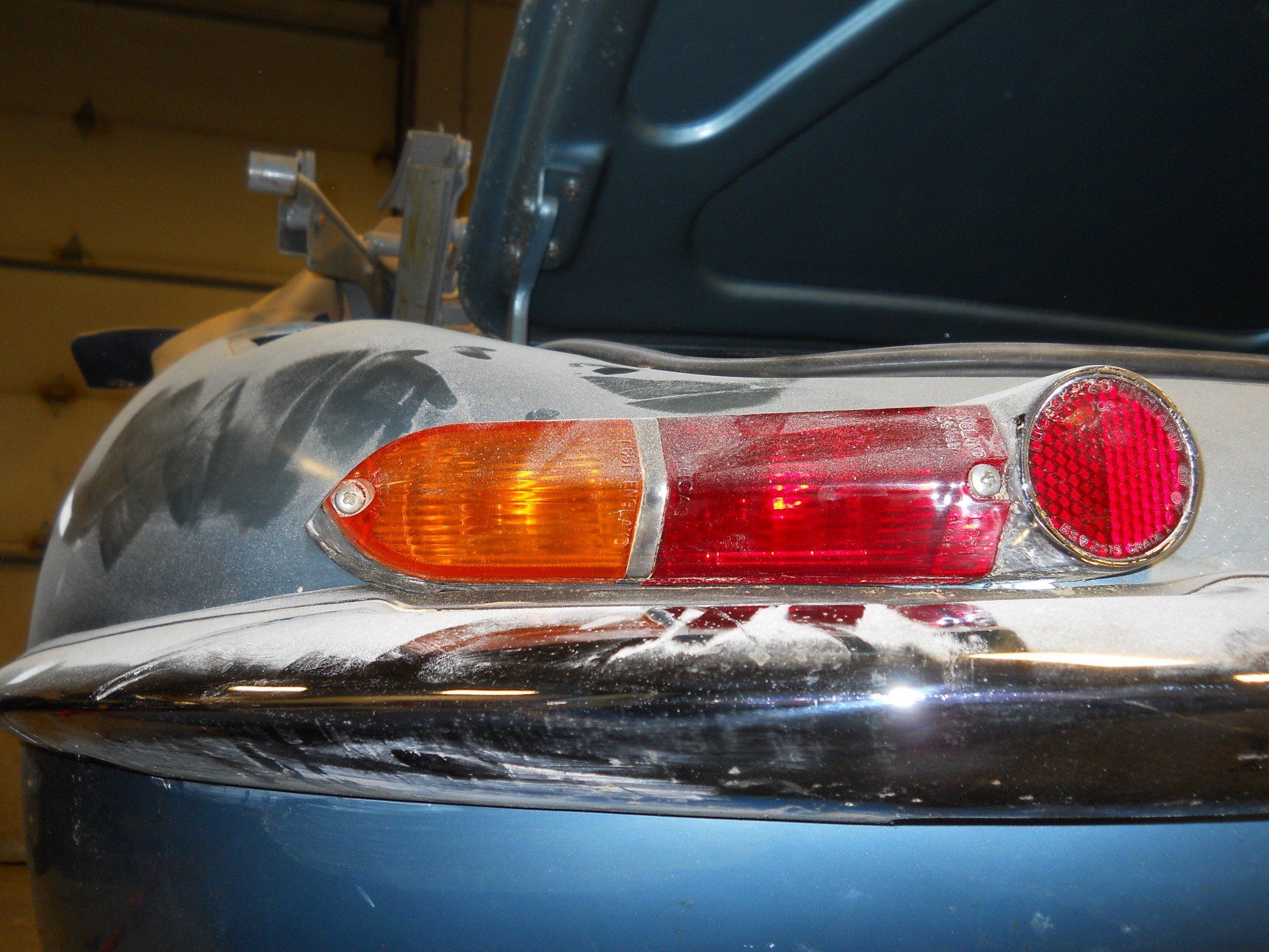 Rear of a blue Jaguar car showing taillights, chrome bumper, and open trunk in a workshop setting.
