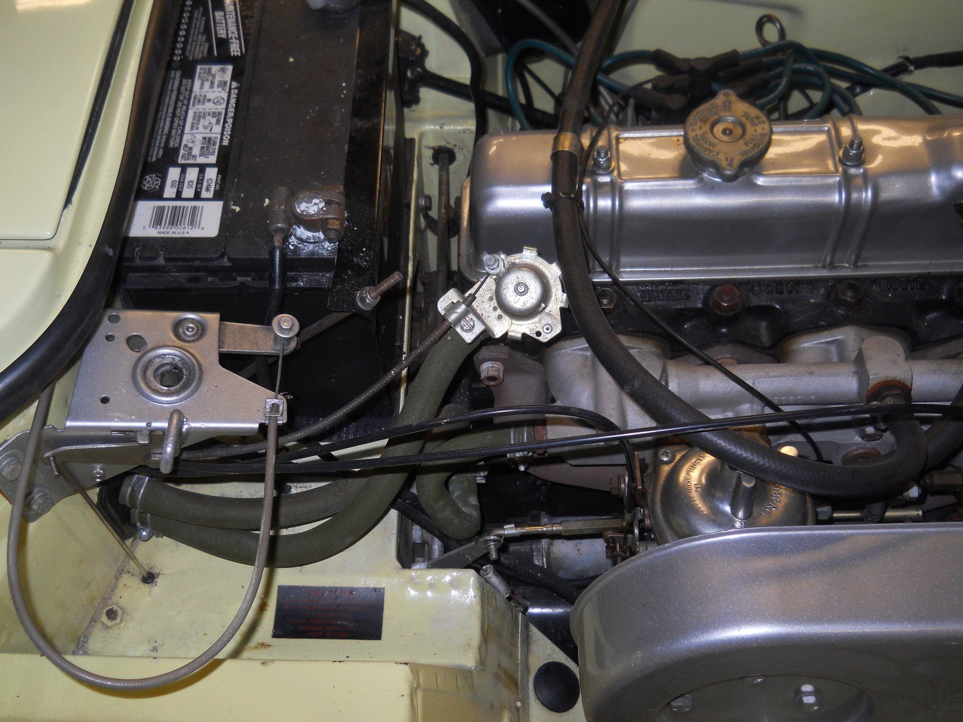 Car engine bay with battery, engine, and various hoses and components.