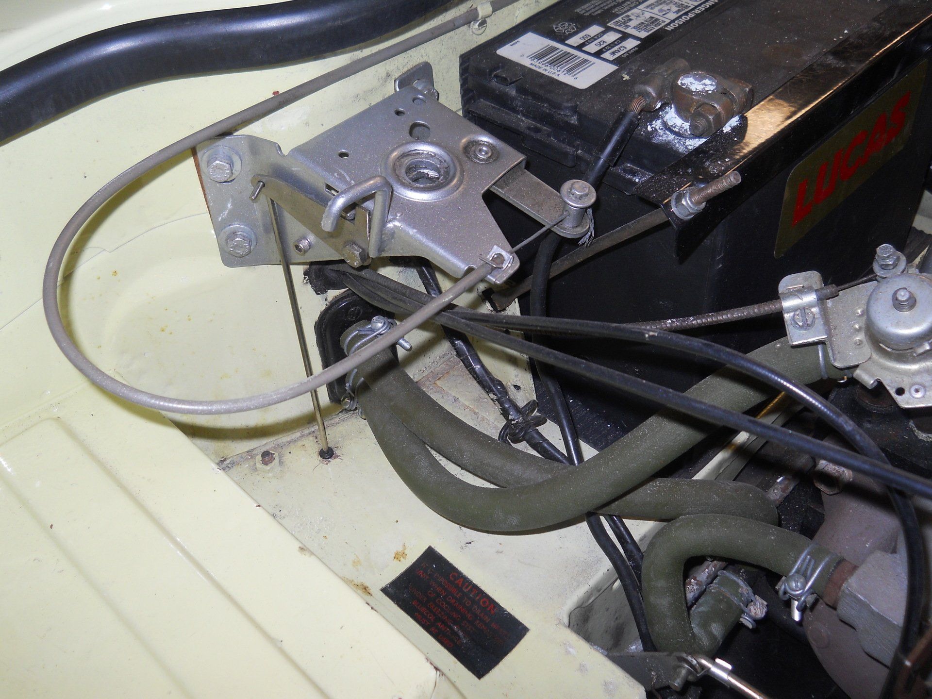 Silver latch and battery in a car engine compartment with black hoses and a yellow surface.