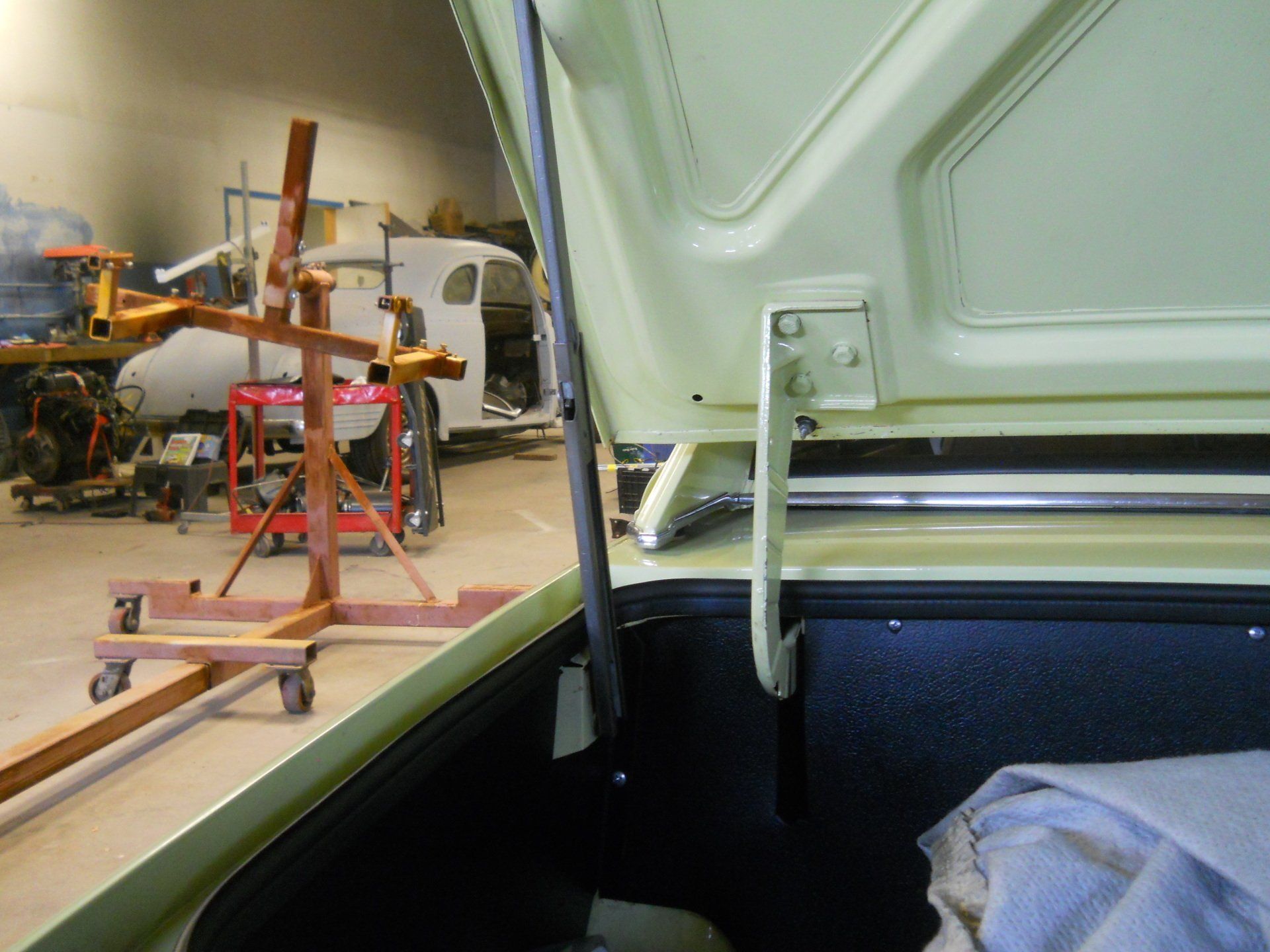Open car trunk, pale yellow, hinges visible. Workshop background with vehicle in progress.