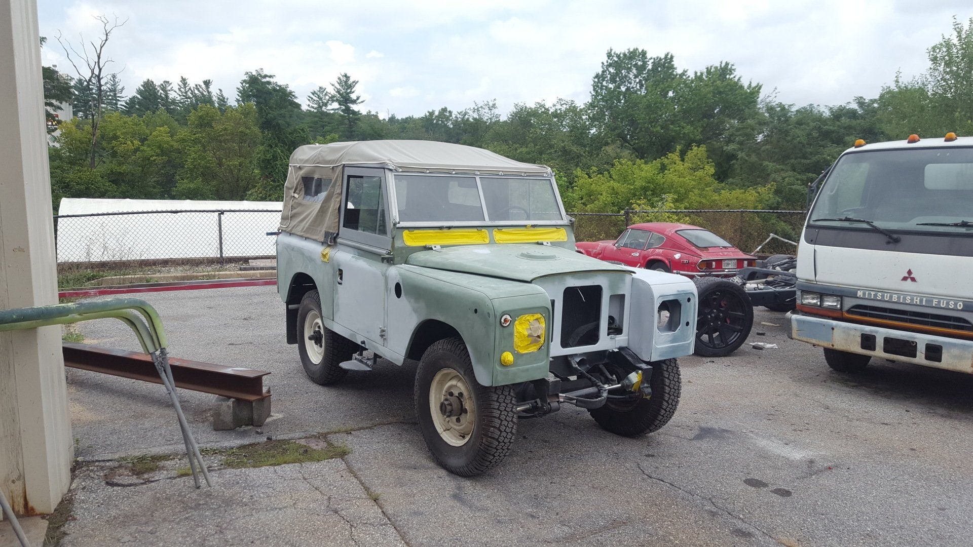 A partly restored green Land Rover with a canvas top, parked outdoors next to a white truck and a red car.