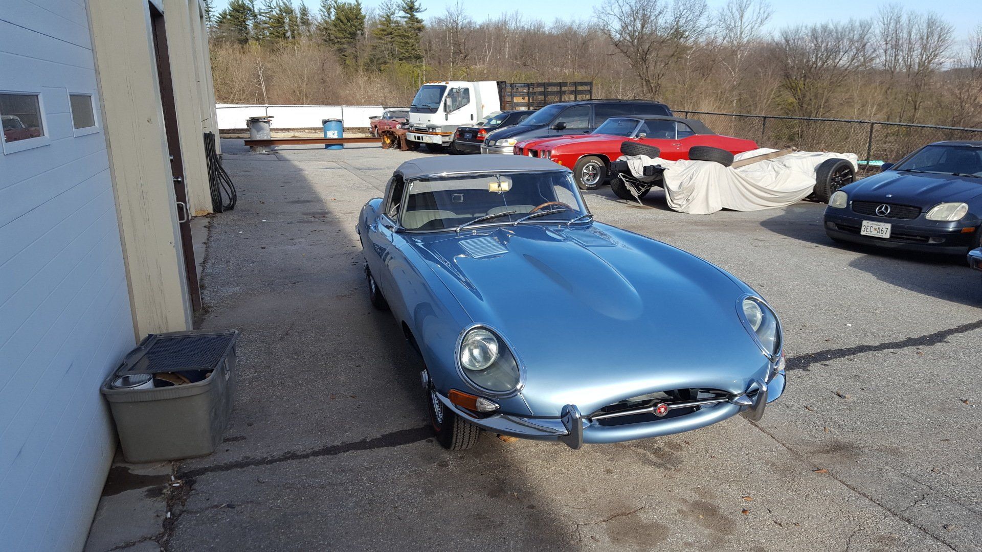Blue Jaguar E-Type convertible parked in front of a garage. Other vehicles visible in the background.