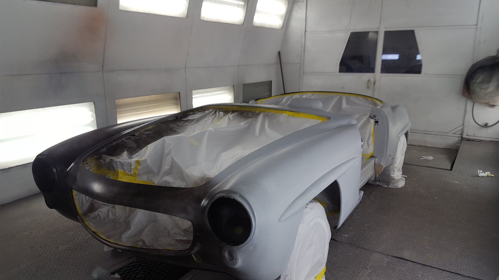 Classic car in a paint booth, partially masked and primed for painting.