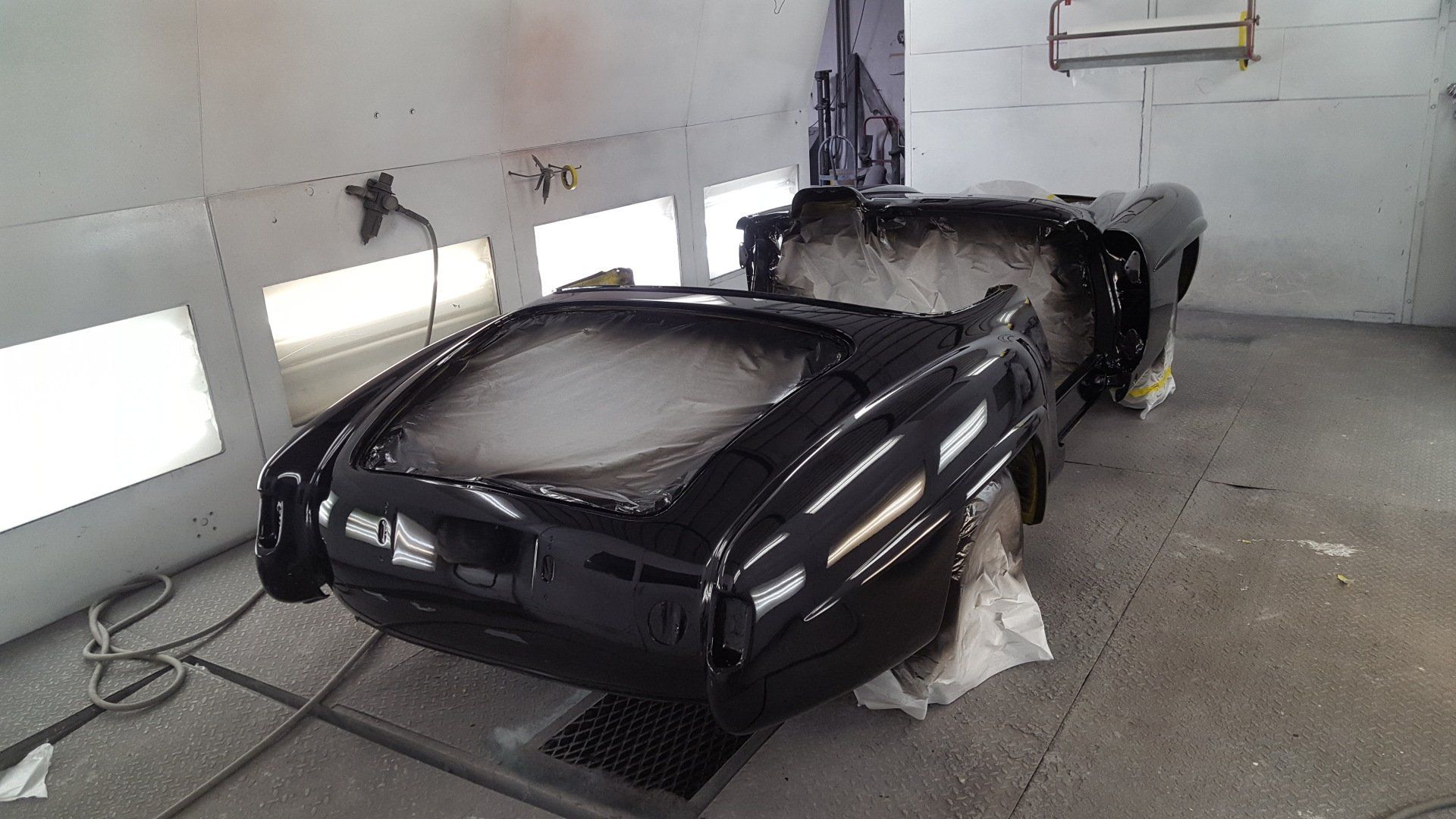 Classic black car in a spray paint booth, partially masked and freshly painted.