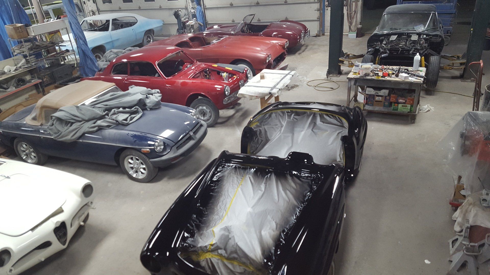 Cars in various stages of restoration inside a workshop; one is being painted black.