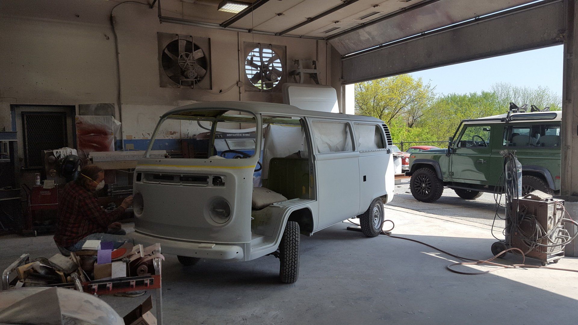 A light-colored van in a garage being worked on, a person welding, and a green SUV parked nearby.