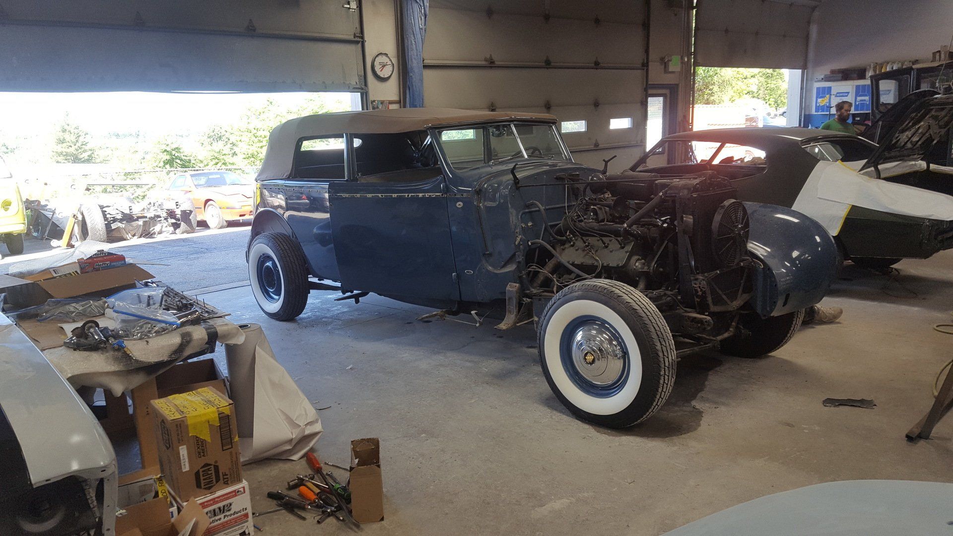A vintage blue car in a workshop, undergoing restoration. It has white-wall tires, missing front body panels.