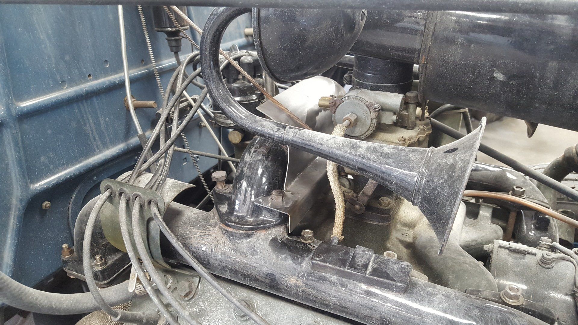 Engine bay of an old vehicle with a trumpet-shaped horn mounted near the carburetor and air filter.