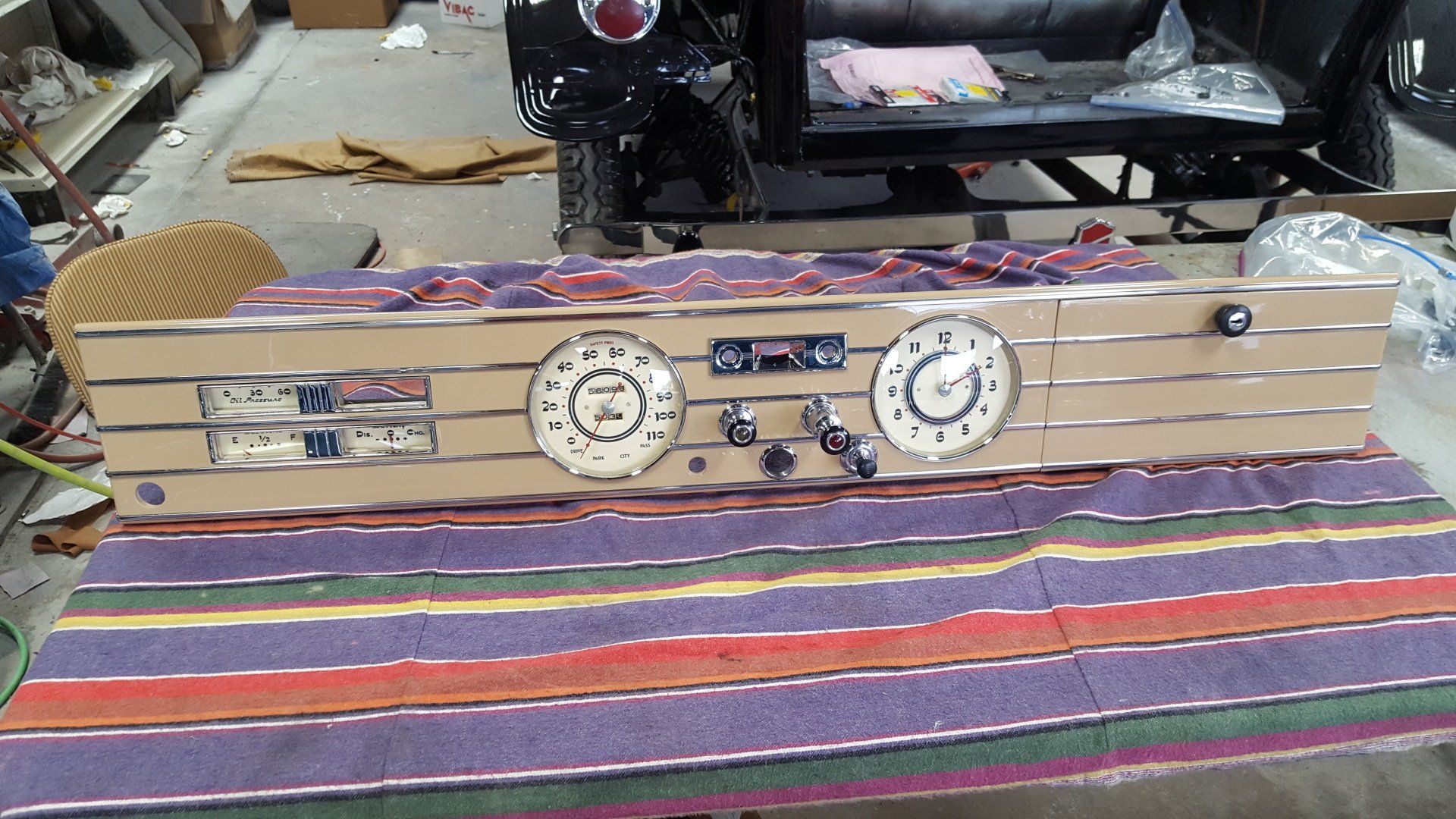 Car dashboard with gauges and knobs, beige panel with silver stripes, resting on a striped fabric.