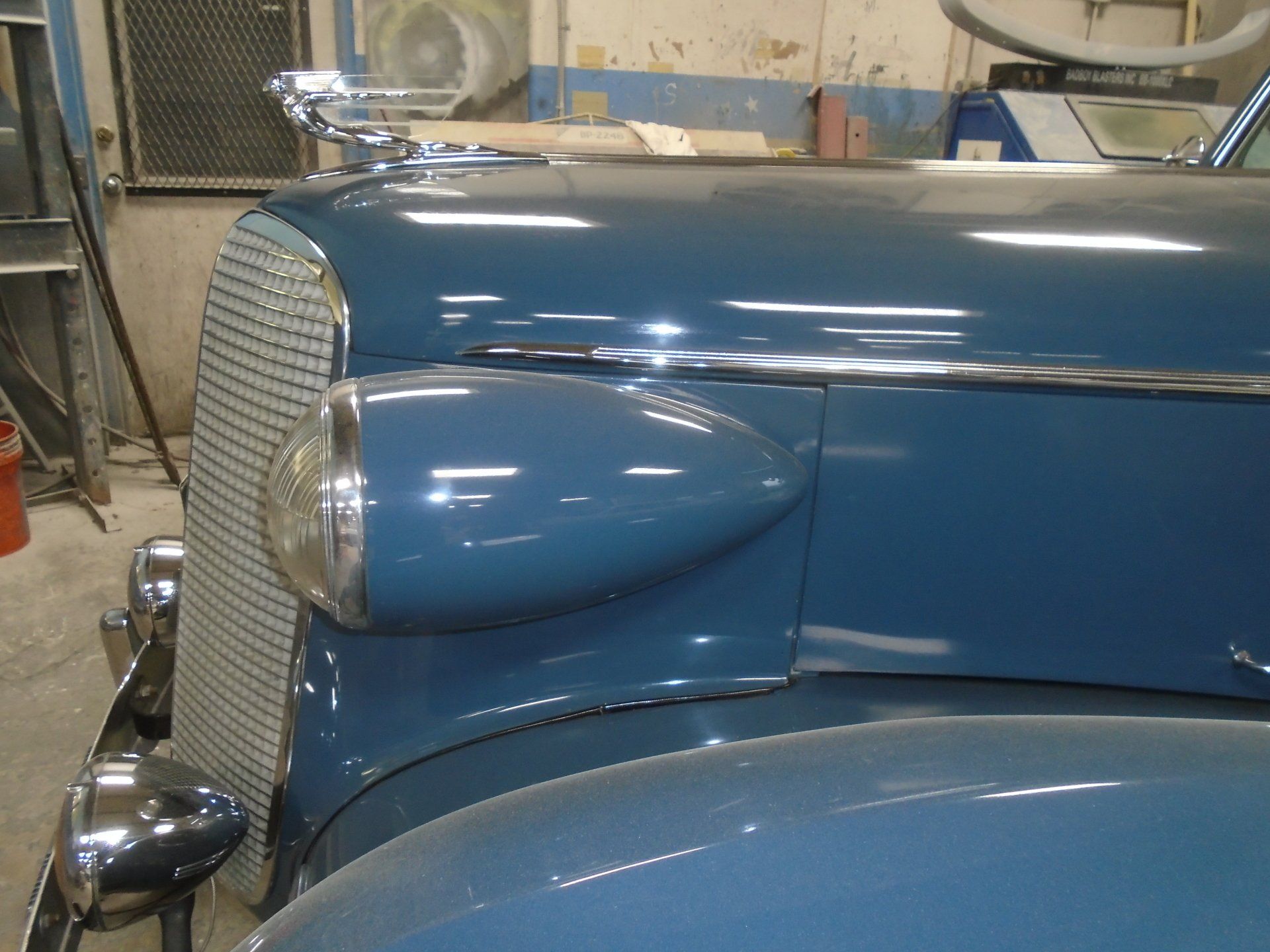 Close-up of a blue vintage car's hood, grille, and headlight, with a chrome emblem.