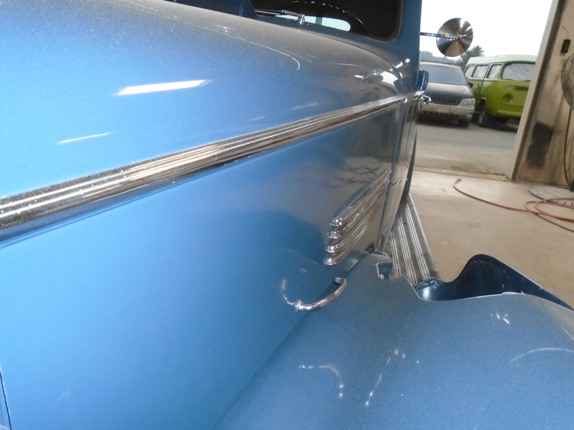 Close-up of a light blue vintage car fender with chrome trim, side mirror, and other vehicles in the background.