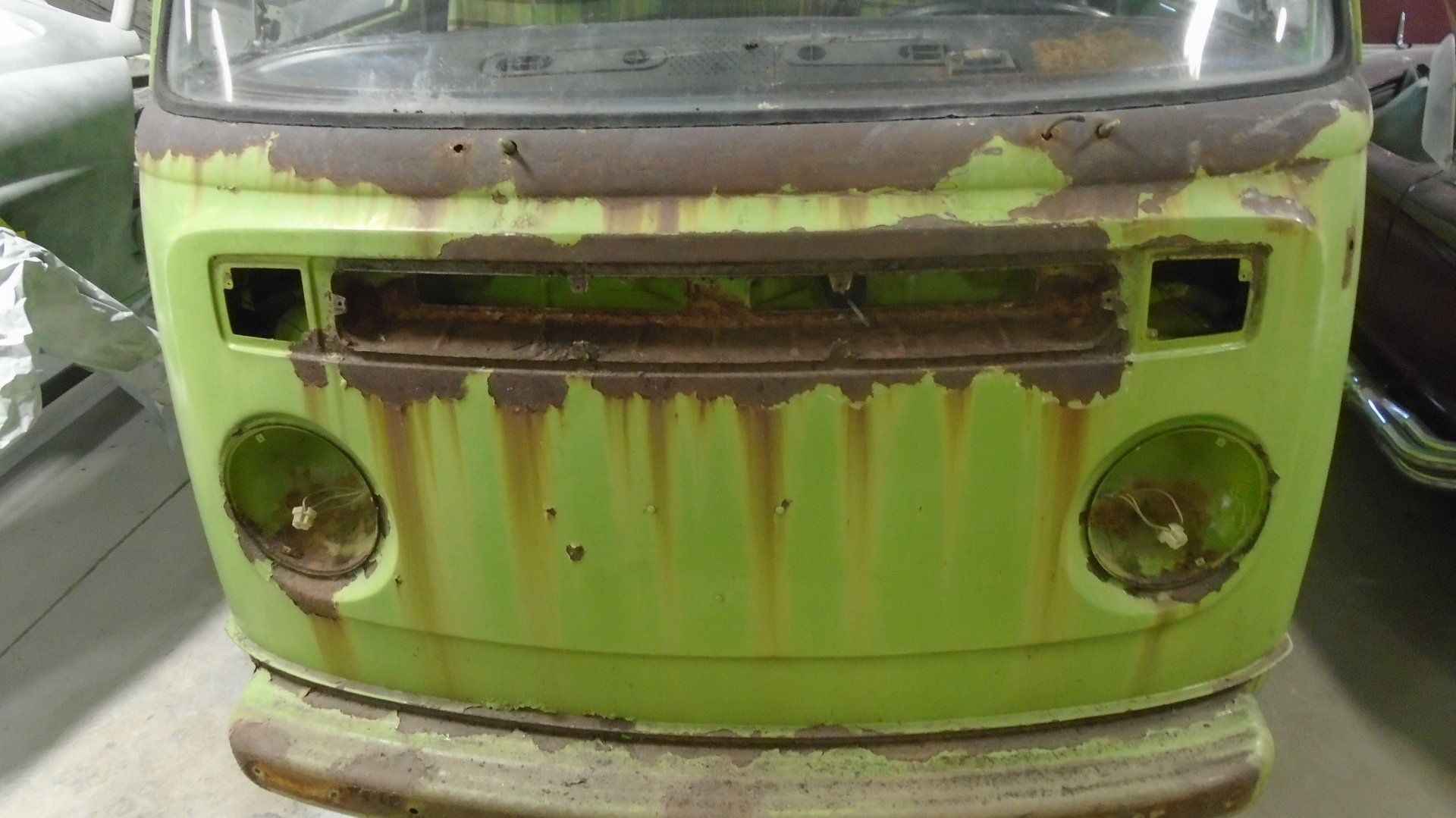 Front of a vintage, lime-green Volkswagen bus with peeling paint, showing rust streaks, empty headlight and grille openings.