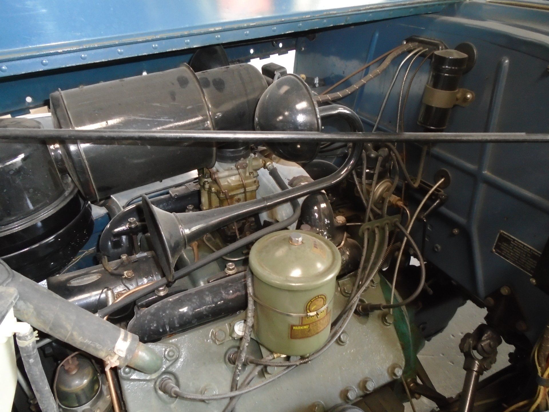 Engine of a vintage blue car, featuring a black air filter, horn, and various mechanical parts.