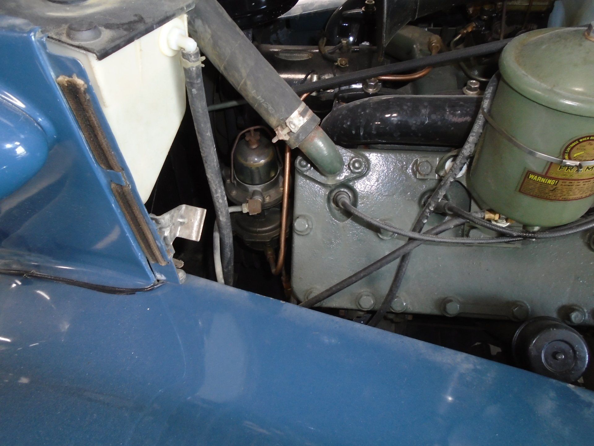 Engine compartment of a blue vintage car, featuring black hoses, a green air filter, and a beige coolant reservoir.