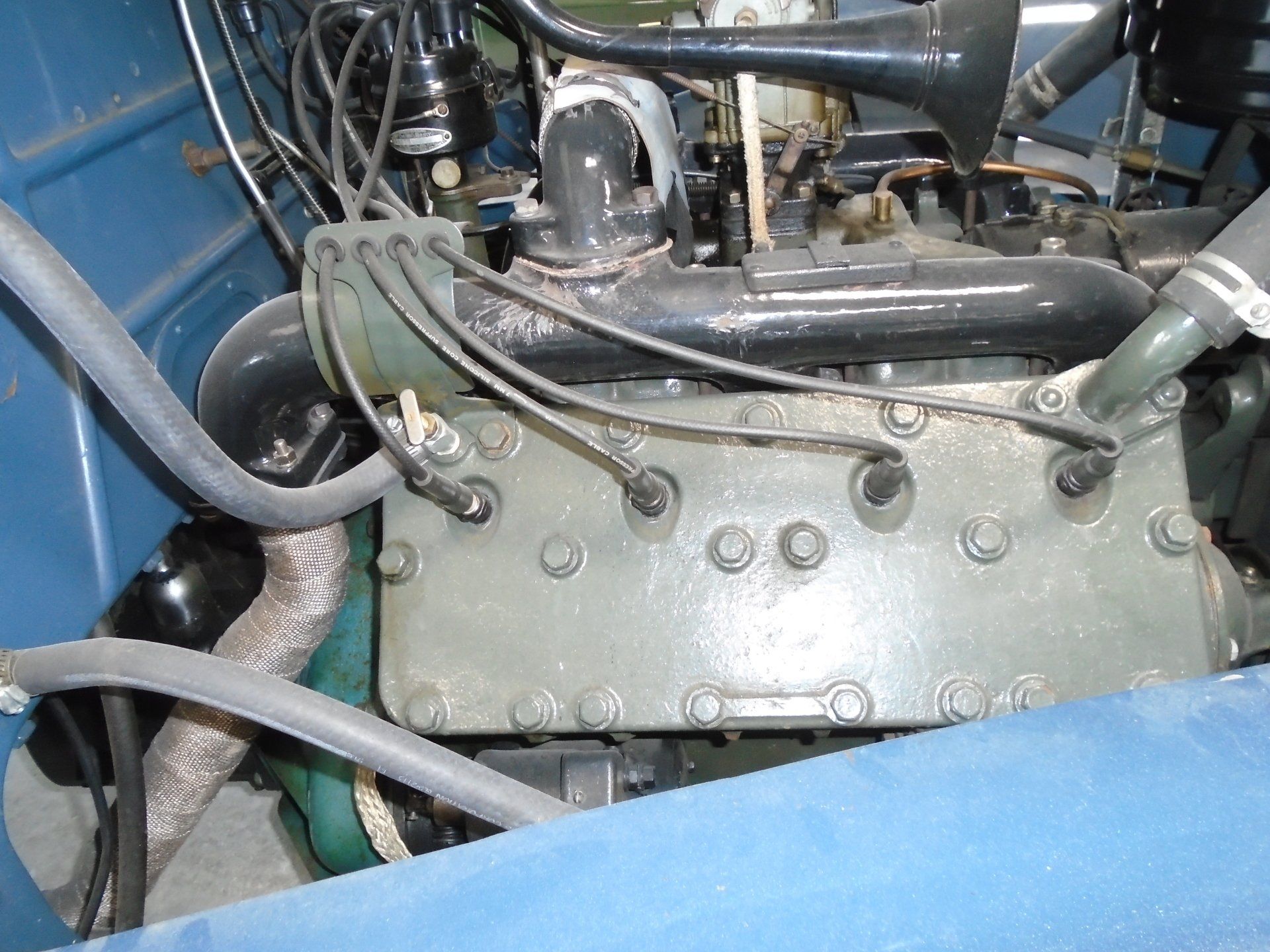 Close-up of a classic car engine with black hoses, gray engine block, and various mechanical parts.