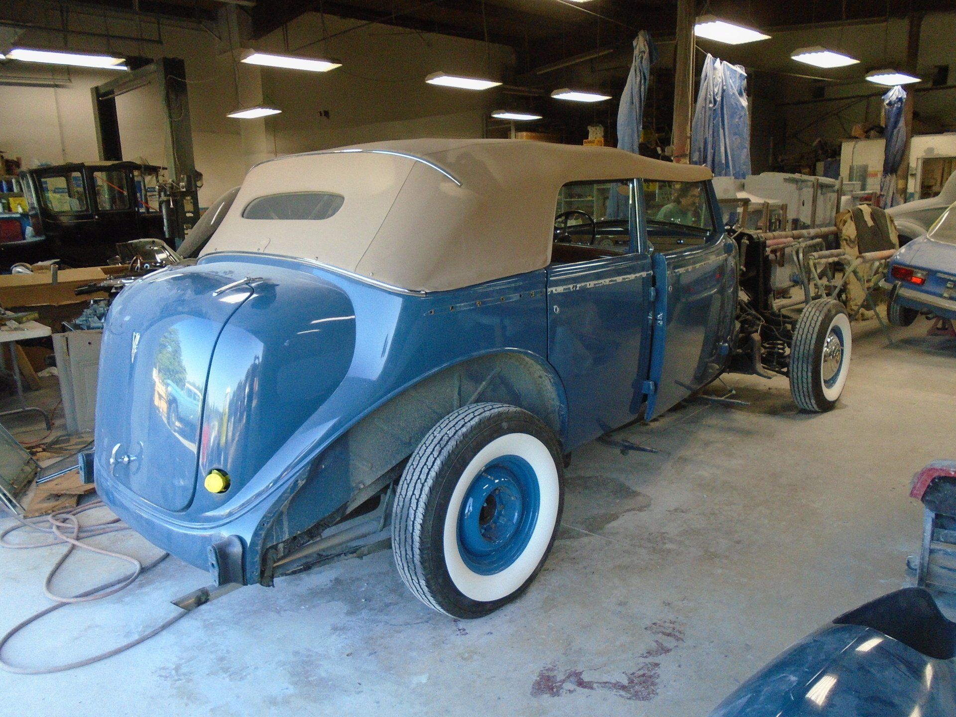Blue vintage car in a garage, restoration in progress, tan convertible top, white wall tires.