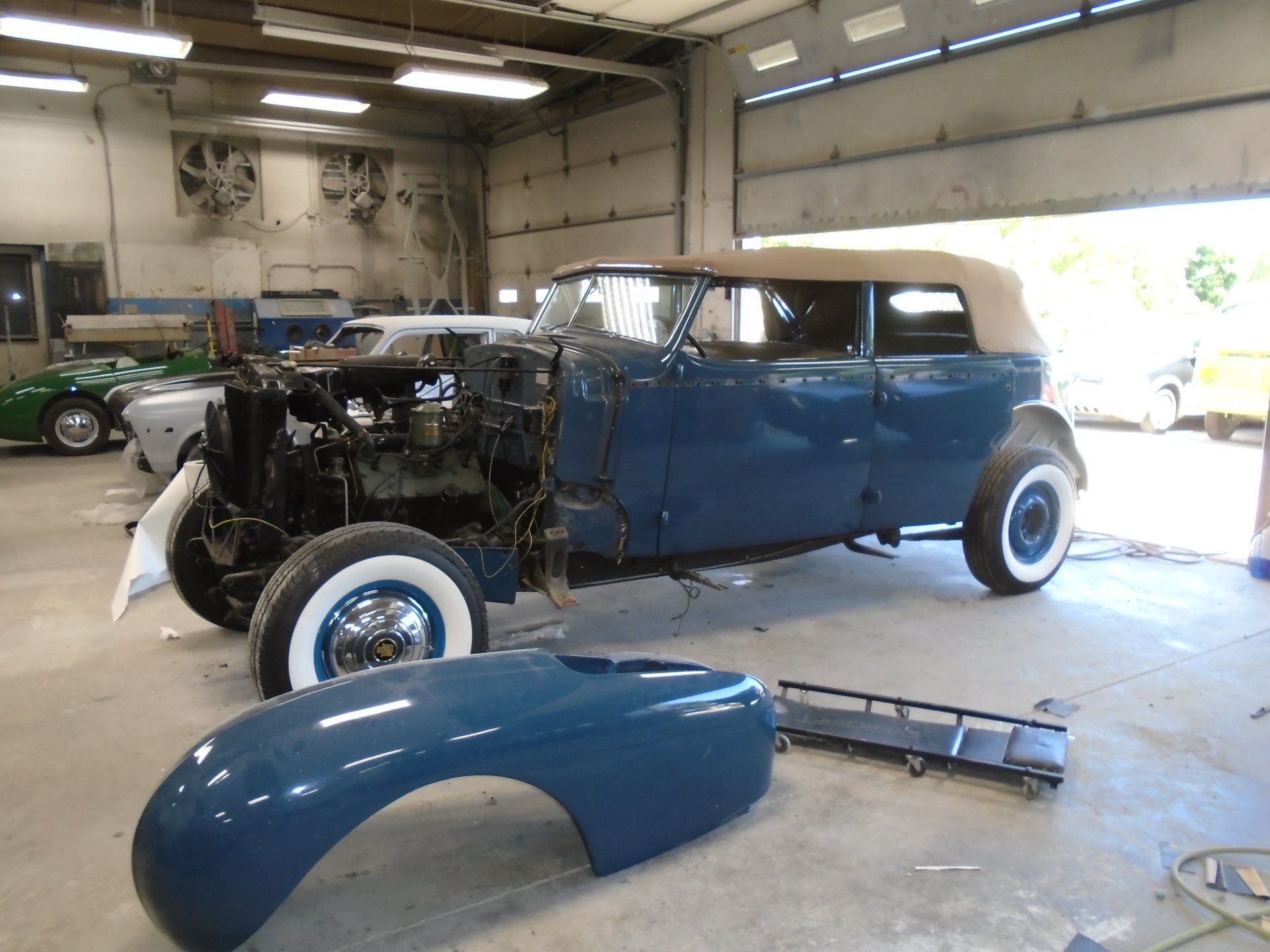 Blue vintage car in a shop, undergoing restoration. Body partially disassembled, white-wall tires.