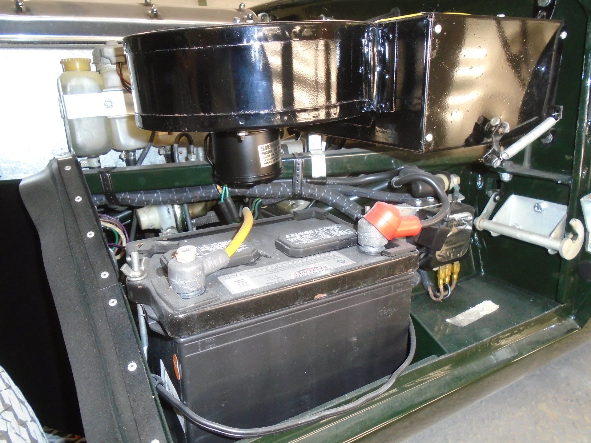 Black car battery in engine compartment, with air filter and fluid containers visible.