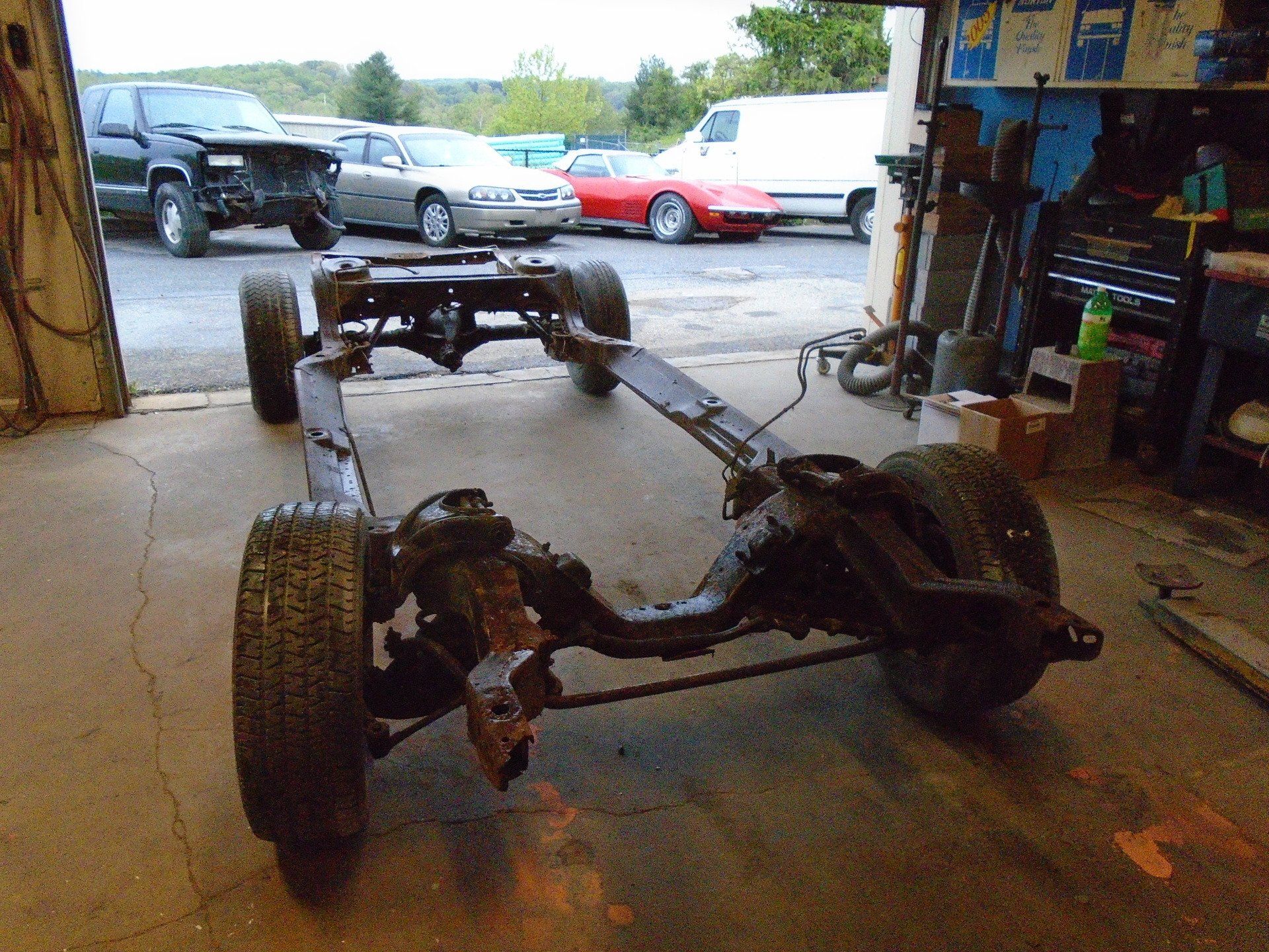 Car chassis on a garage floor with wheels attached, other vehicles visible in the background.