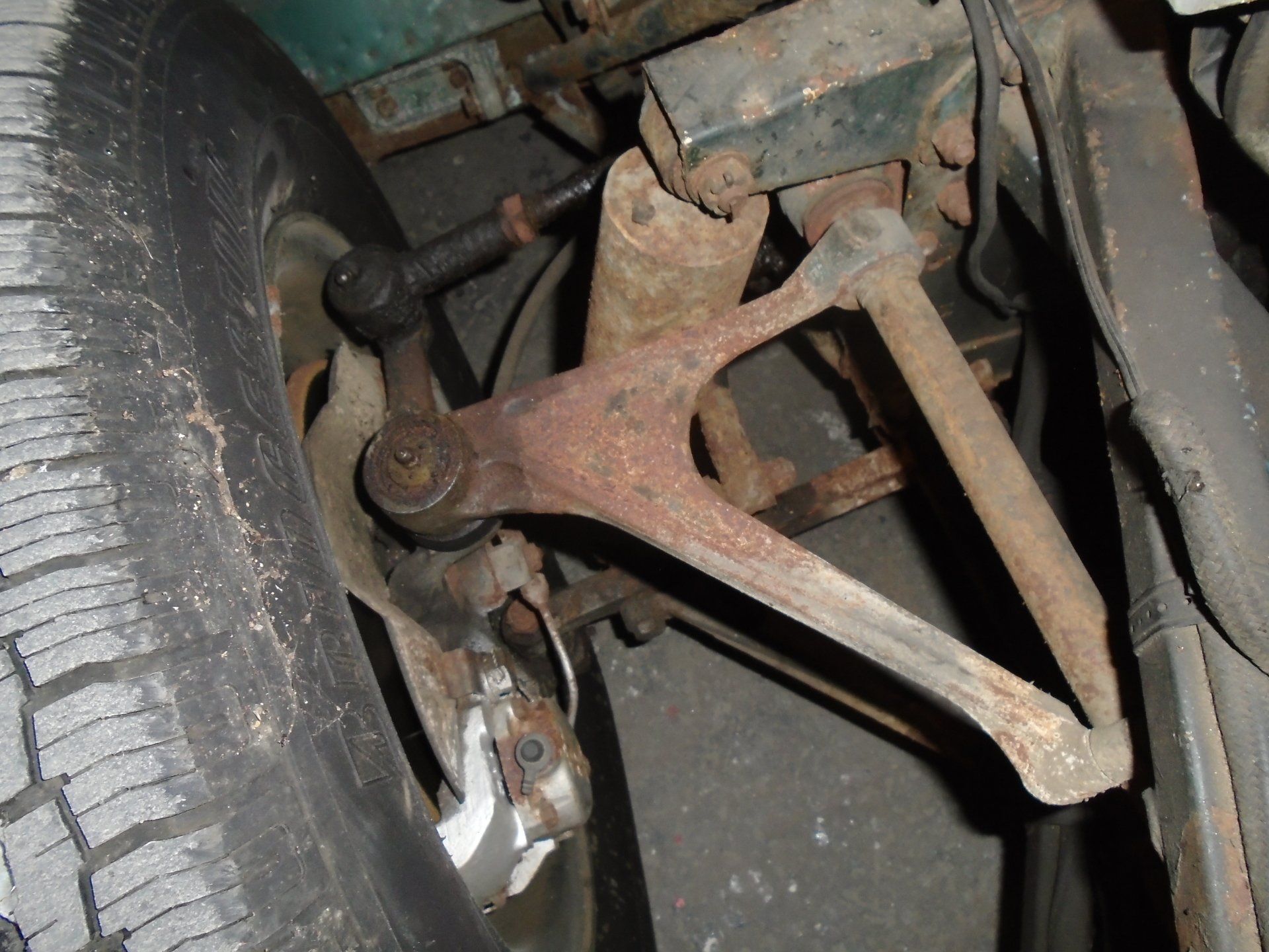 Rusty car suspension components, including wishbone and tie rod, viewed from beneath the wheel.