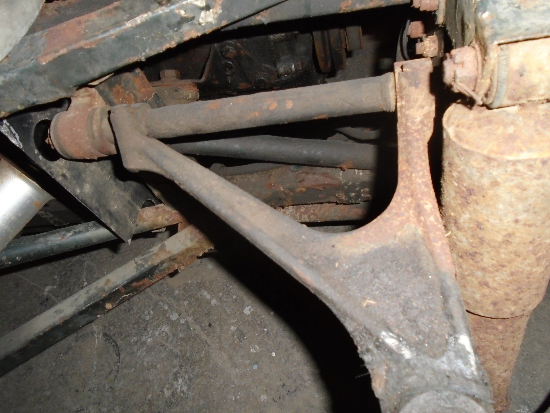 Rusty car suspension components. An angled arm connects to a horizontal bar, shock absorber visible.