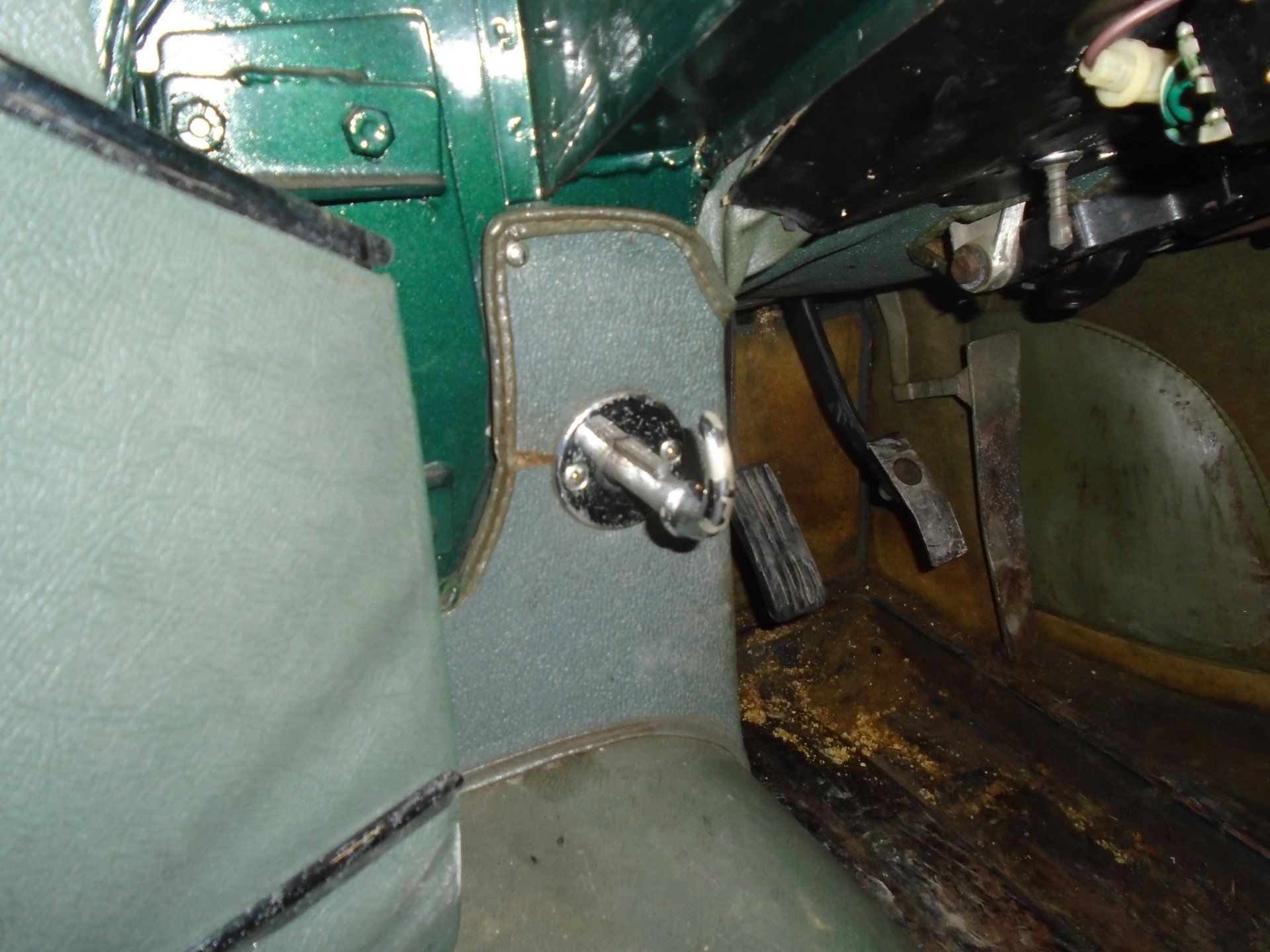 Interior of a green car, showing pedals, a footrest, and a metal hook on the side.