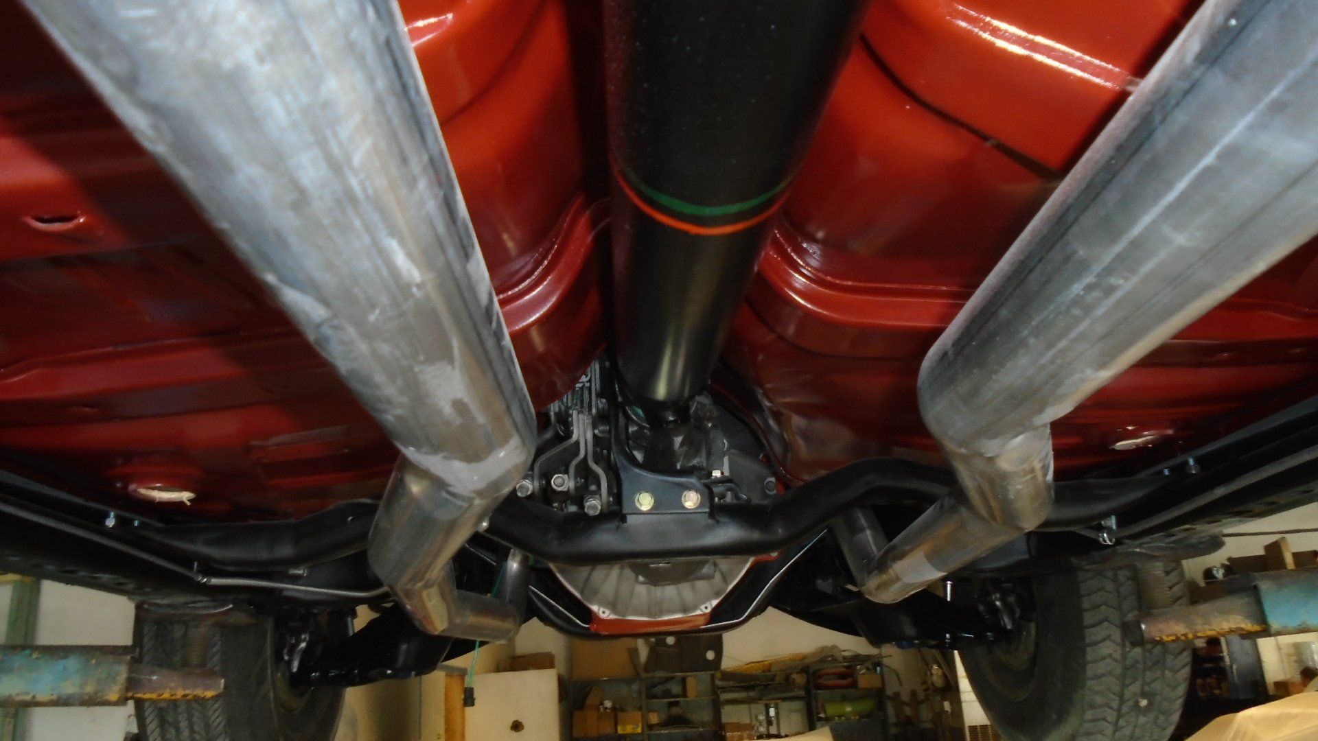 Underside of a classic car showing exhaust pipes, a driveshaft, and rear axle, all against a red floor.