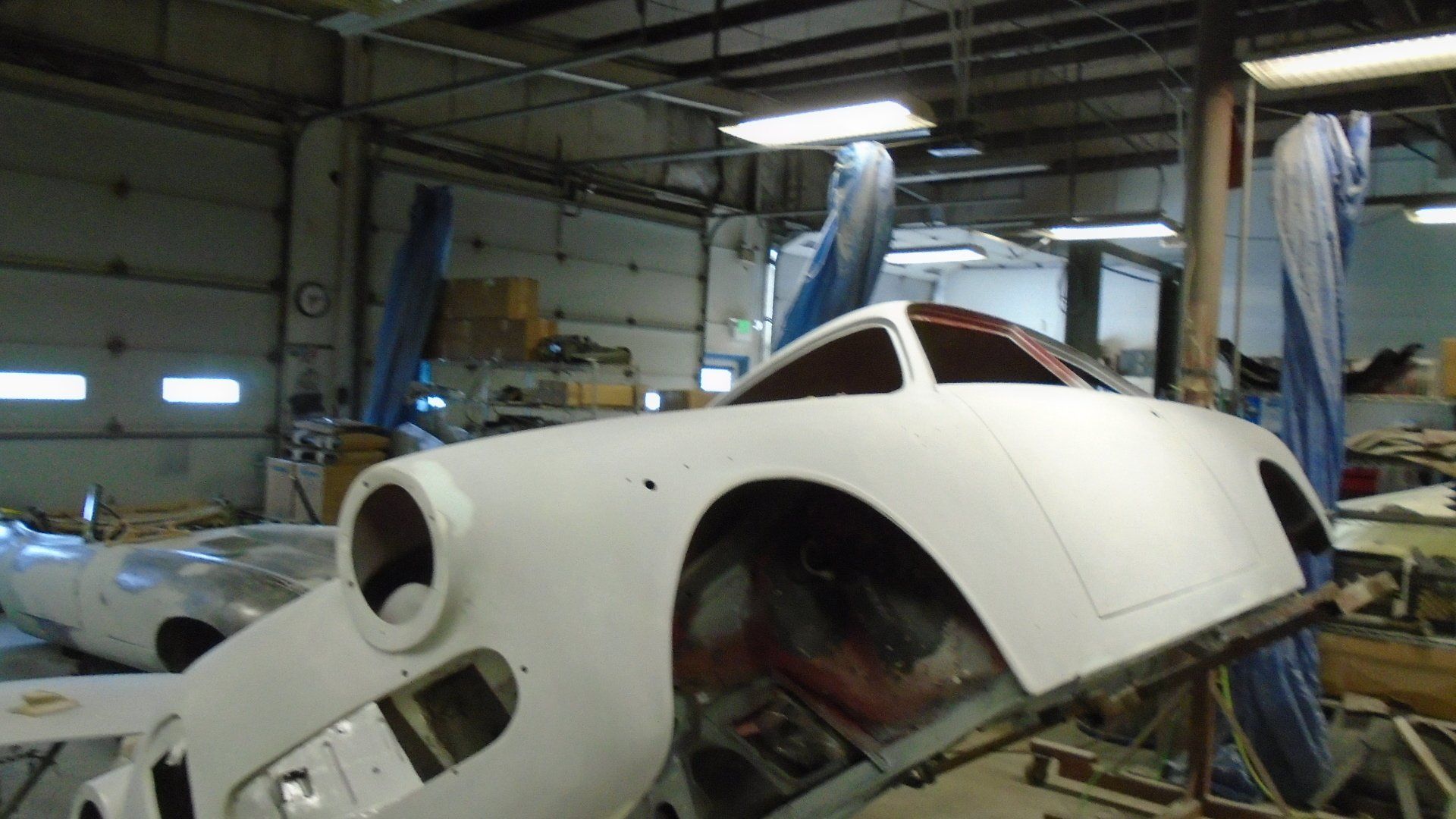 White car body in a workshop, window frame, headlamps visible, covered with blue sheets.