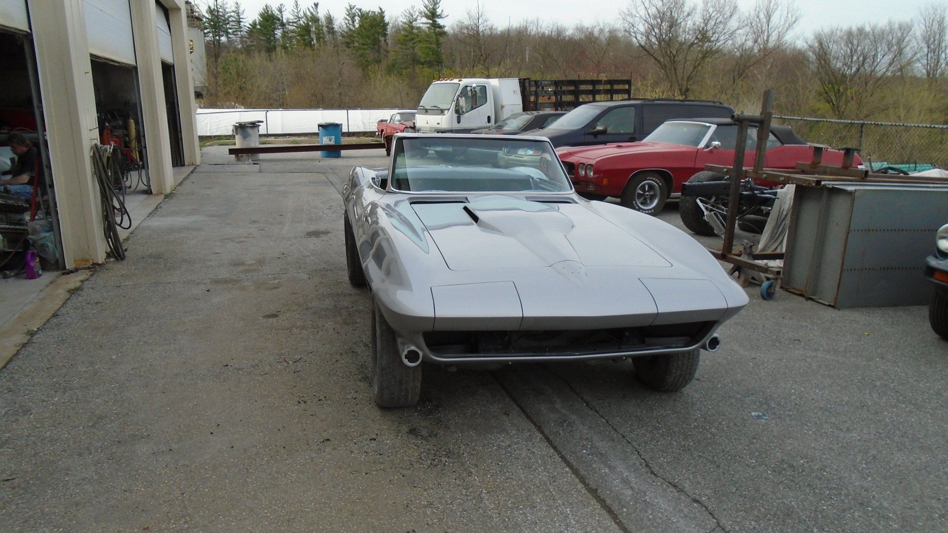 Silver convertible Corvette in a garage, other vehicles in background.