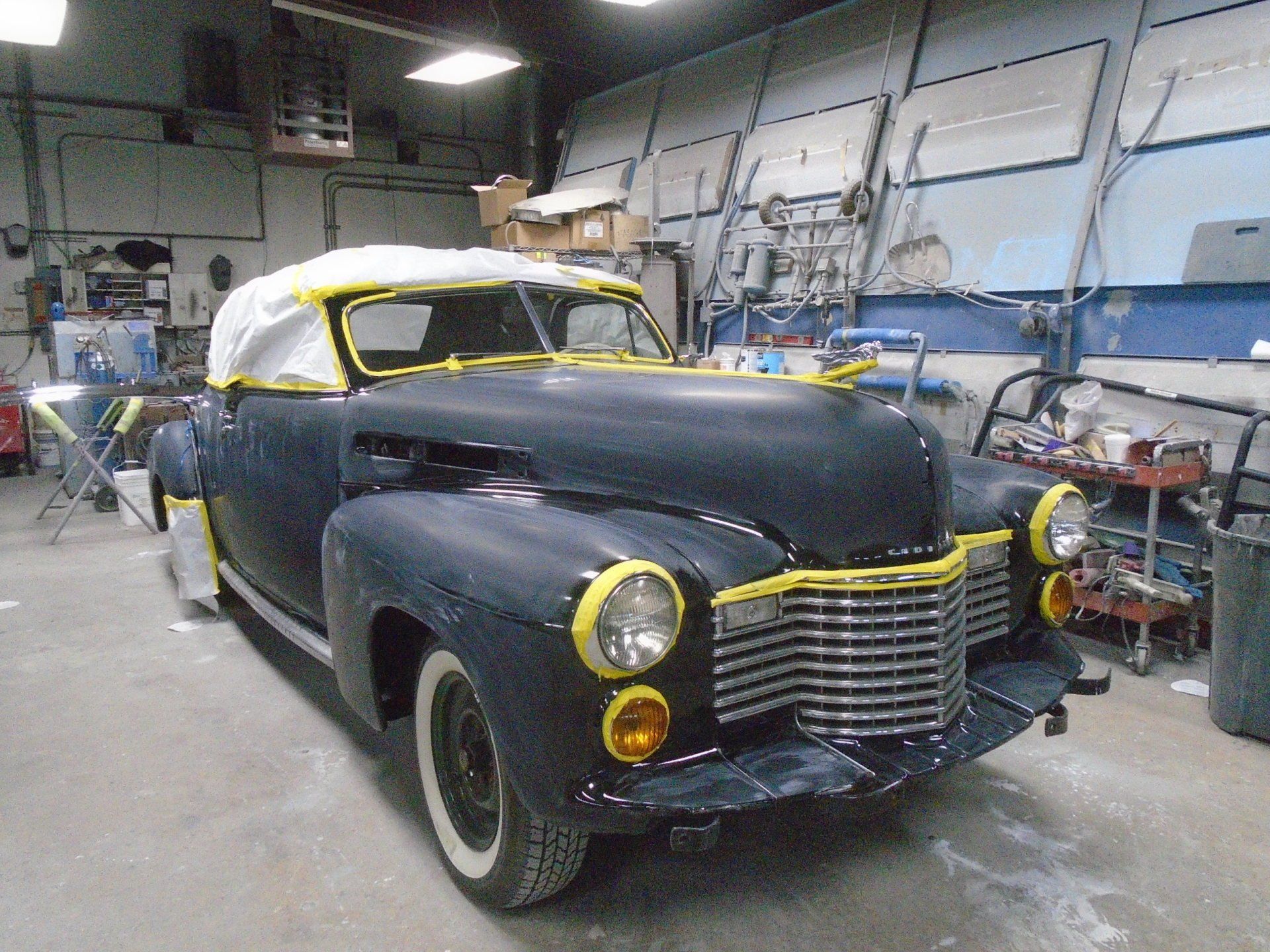 Black classic car in a workshop, masked for painting. White tires, yellow tape, and a white covering.