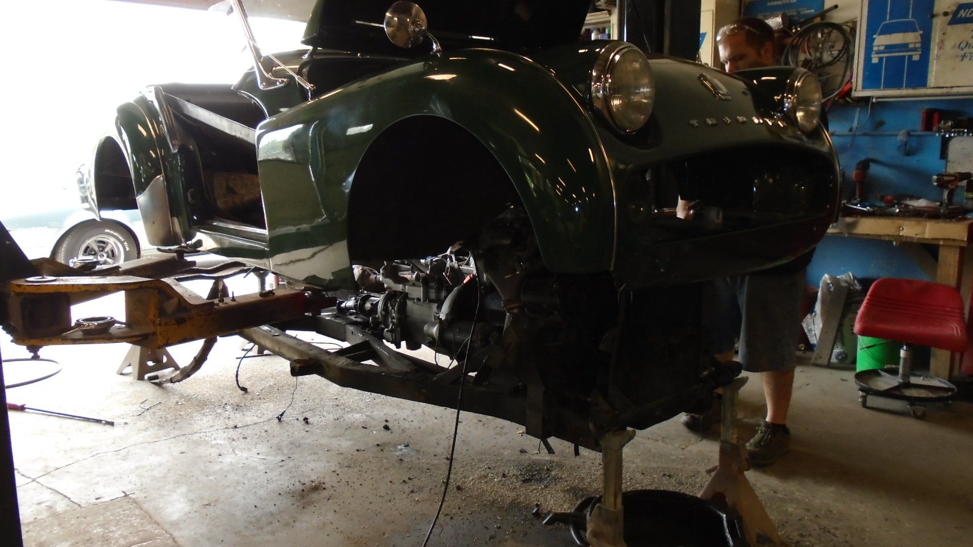 Green classic car in a workshop, on a jack, with a person working on it.
