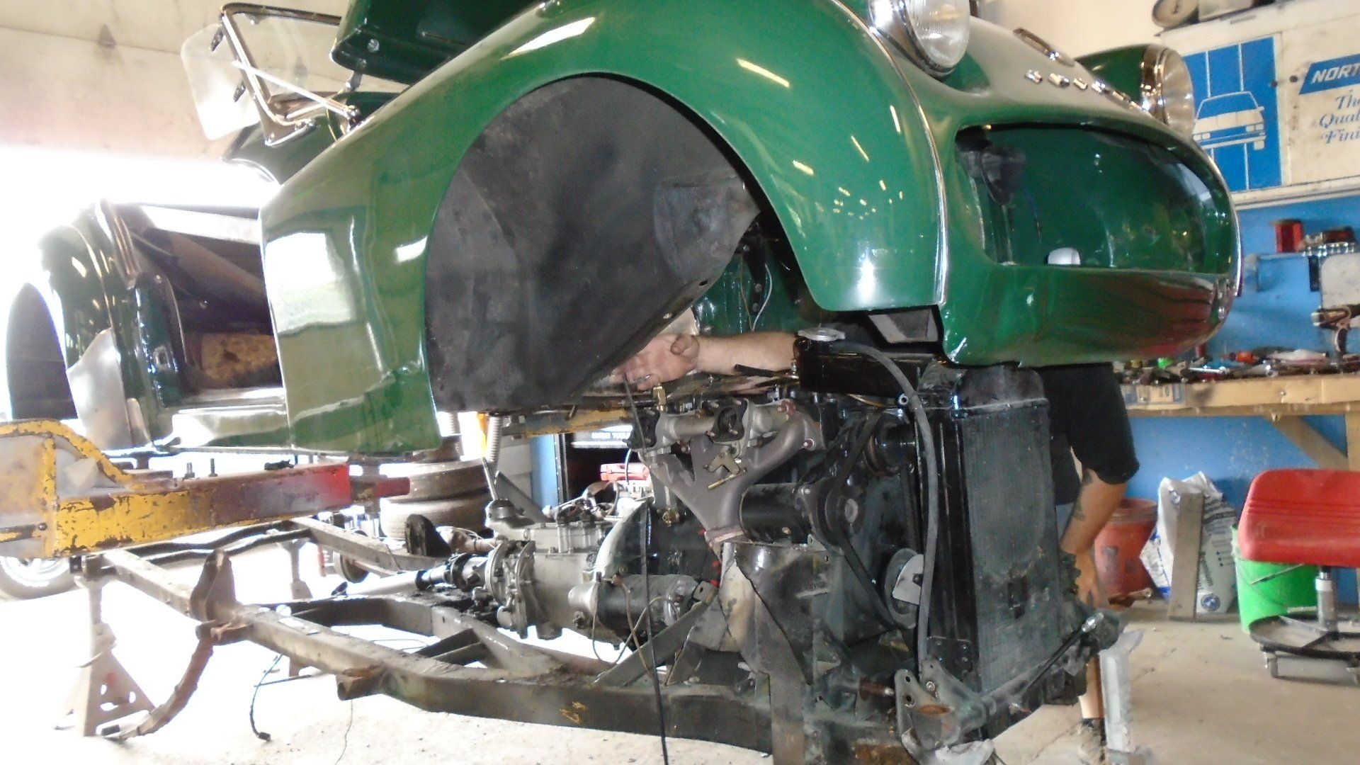 Green vintage car body lifted above engine, man working inside the shop.