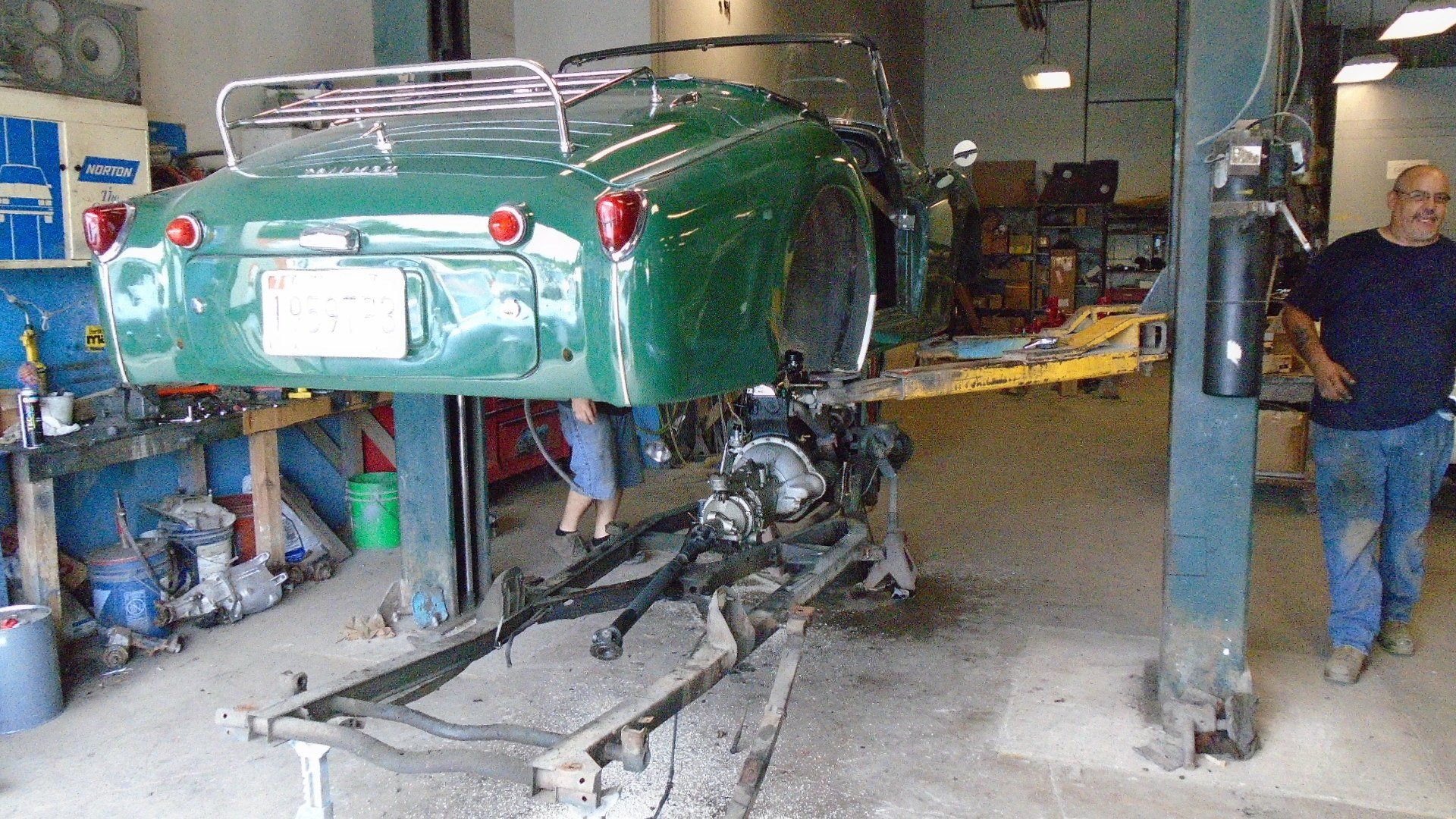 Green convertible car raised in a repair shop. A man stands beside it.