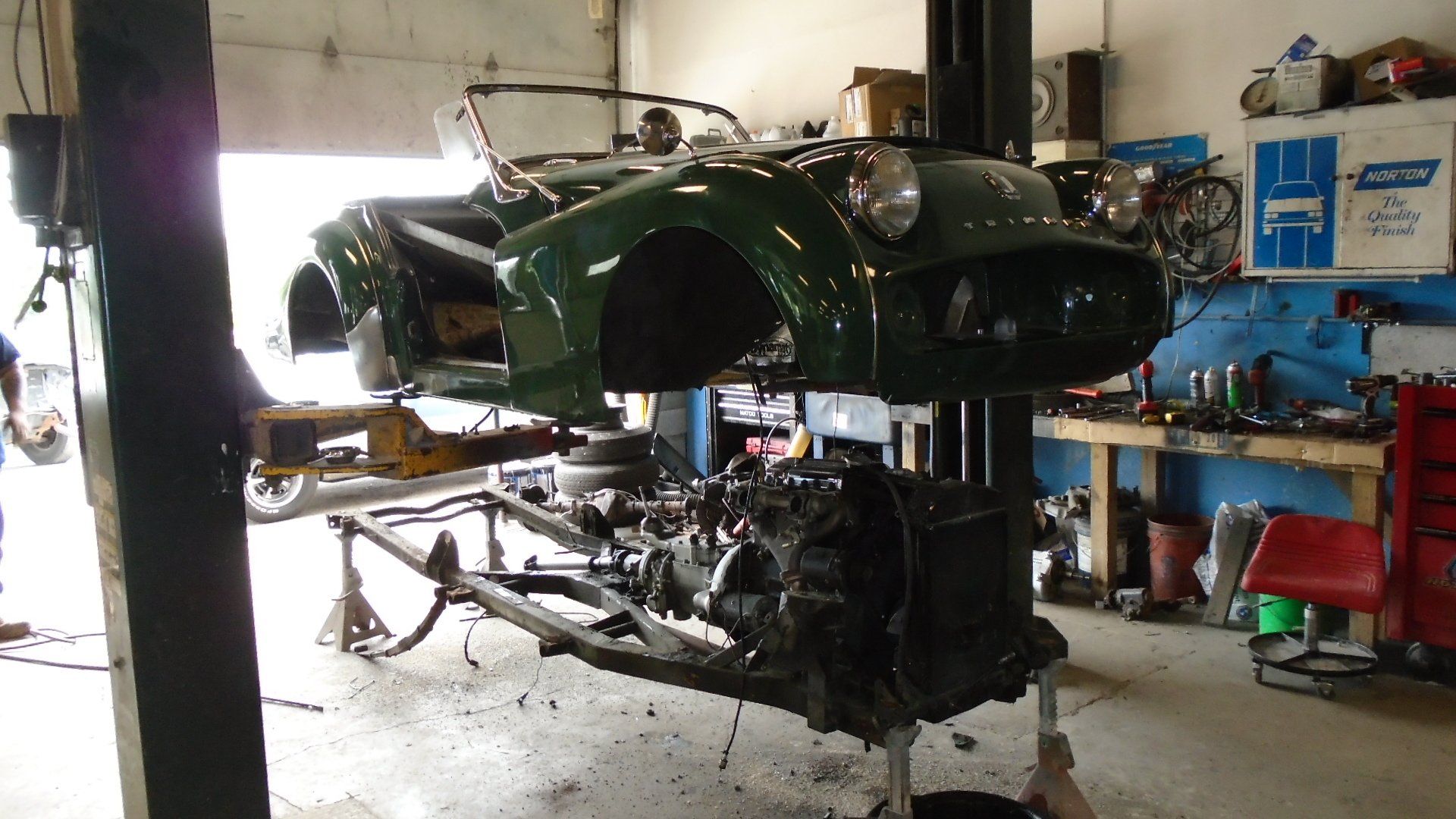 Green Triumph TR3 sports car being worked on in a garage, body raised above frame on a lift.