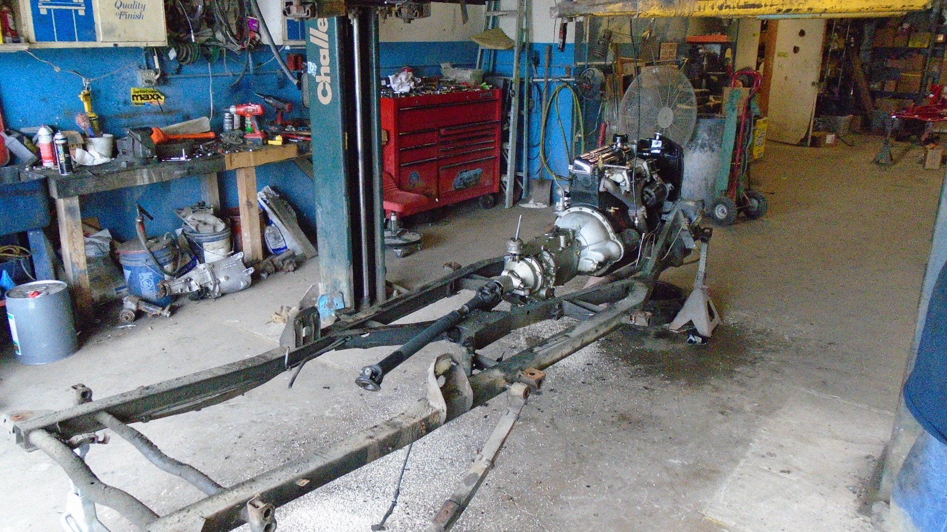 Car chassis and engine in a garage; tools and equipment visible; blue walls.