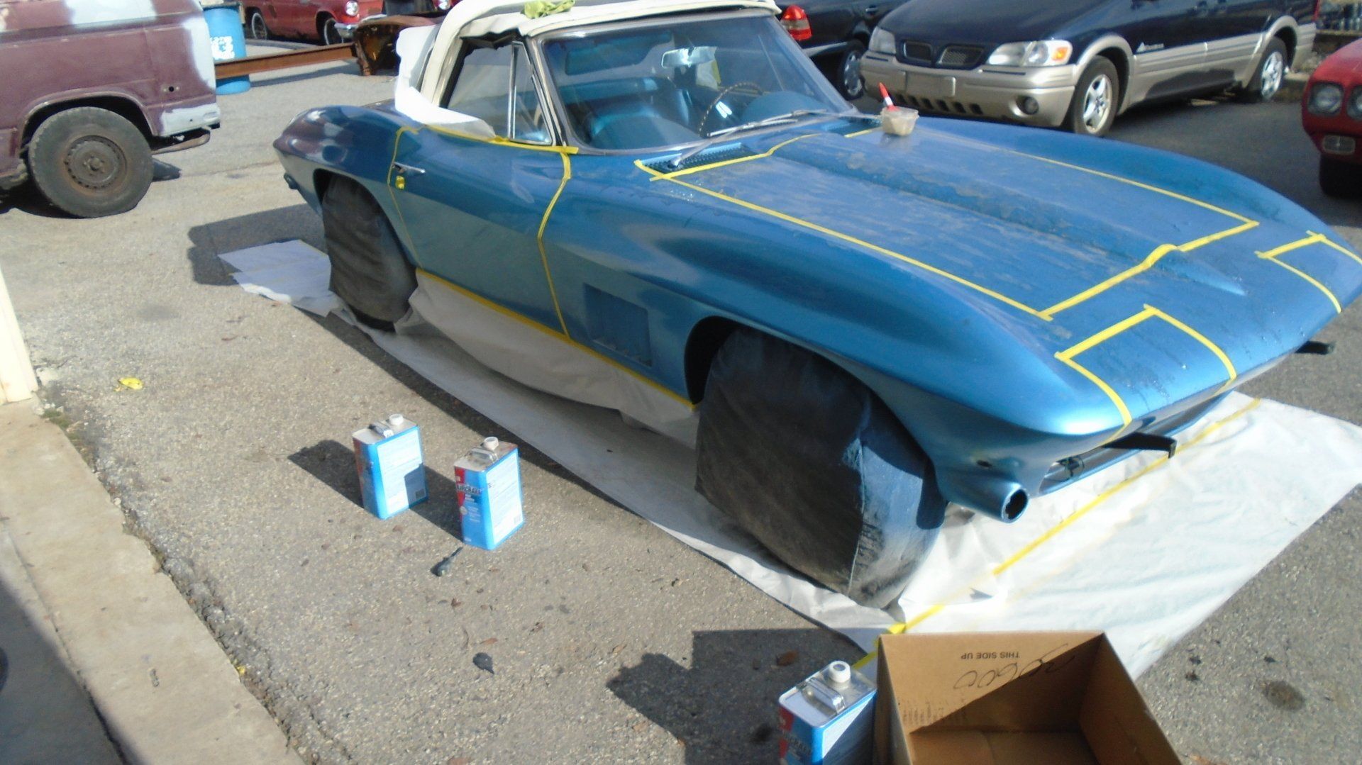 Blue convertible car being painted outdoors, masked with yellow tape, on a white sheet.
